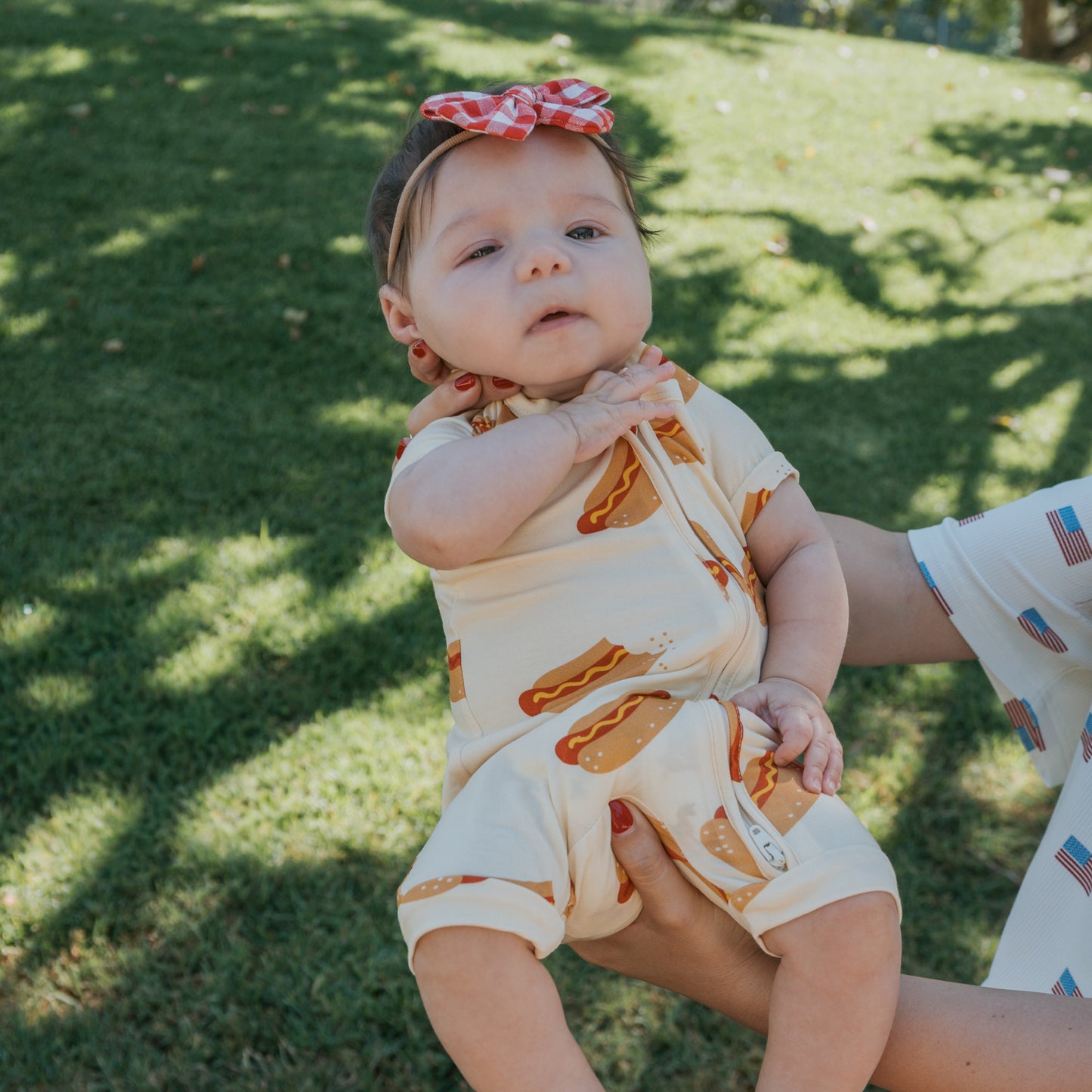A baby wears the forever french baby Shortie Bamboo Zip | Hot Dogs romper and a red gingham headband while being held on a grassy lawn, sunlight filtering through trees—perfect for cute, hypoallergenic style.