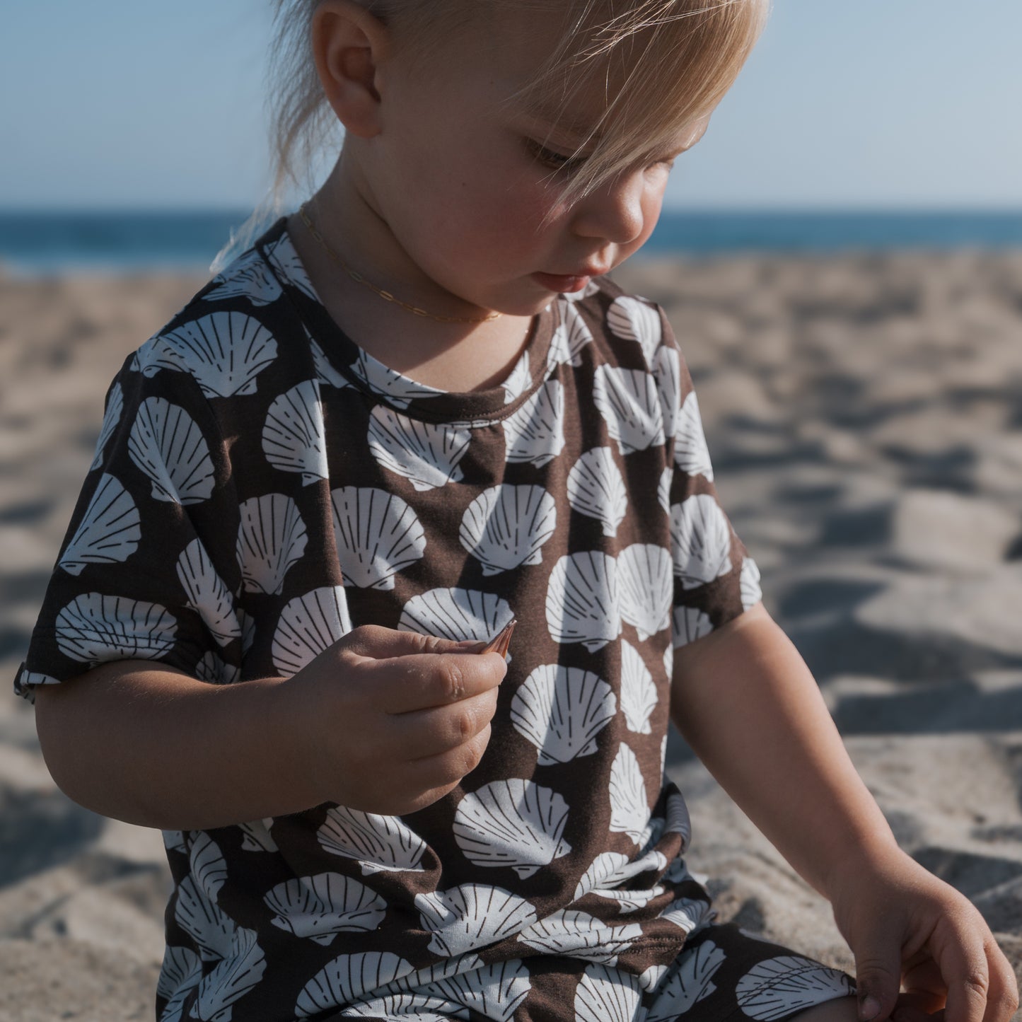 A young child wears the forever french baby Bamboo Short Set | Seashells and sits on a sandy beach, playing with something in their hand as the sea and blue sky create a beautiful backdrop.