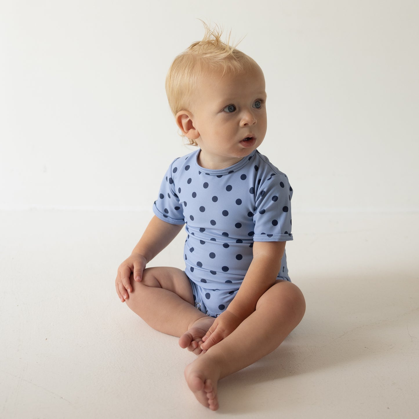 A baby with light hair sits on the floor in a forever french baby Child Shortie Swimsuit | Sully, looking off to the side against a plain white background.