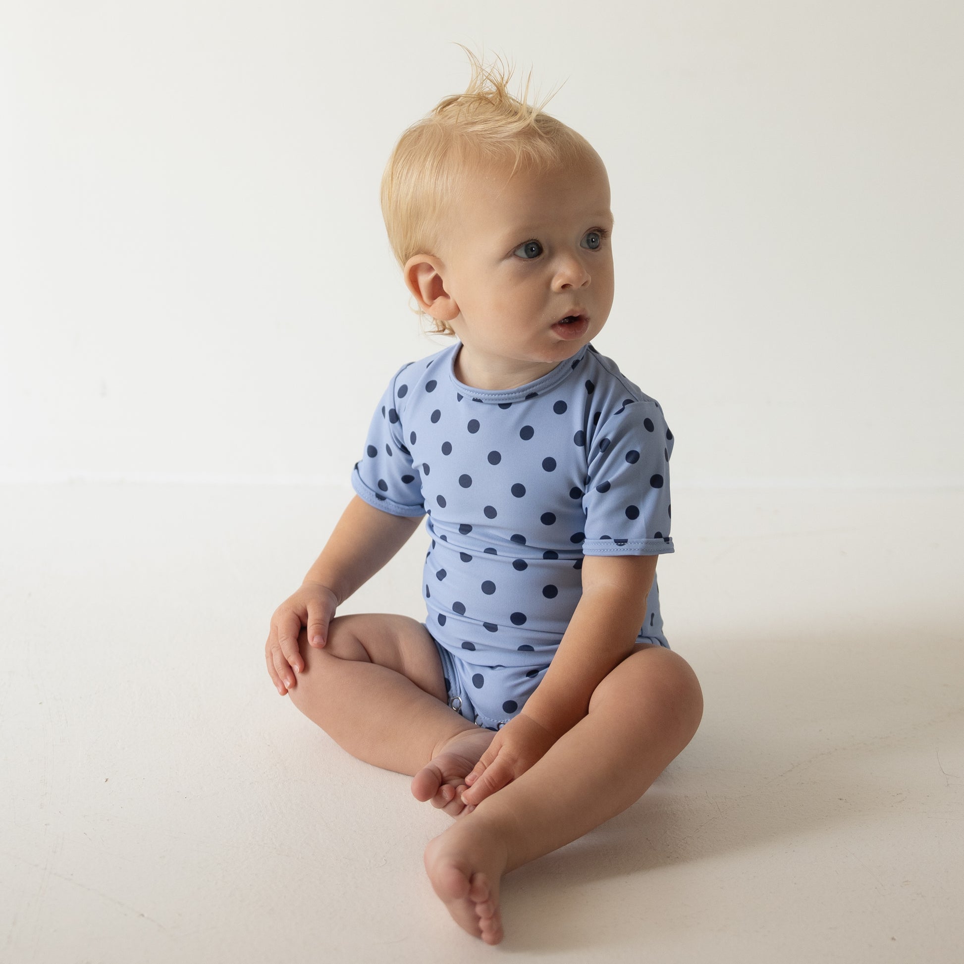 A baby with light hair sits on the floor in a forever french baby Child Shortie Swimsuit | Sully, looking off to the side against a plain white background.
