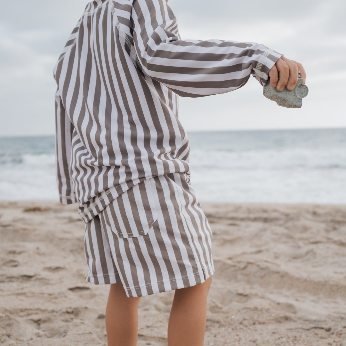 A child stands on a sandy beach facing the ocean, wearing forever french baby’s Child Board Short in Olive Stripe. The cloudy sky and sea are visible in the background as they hold a small rock.