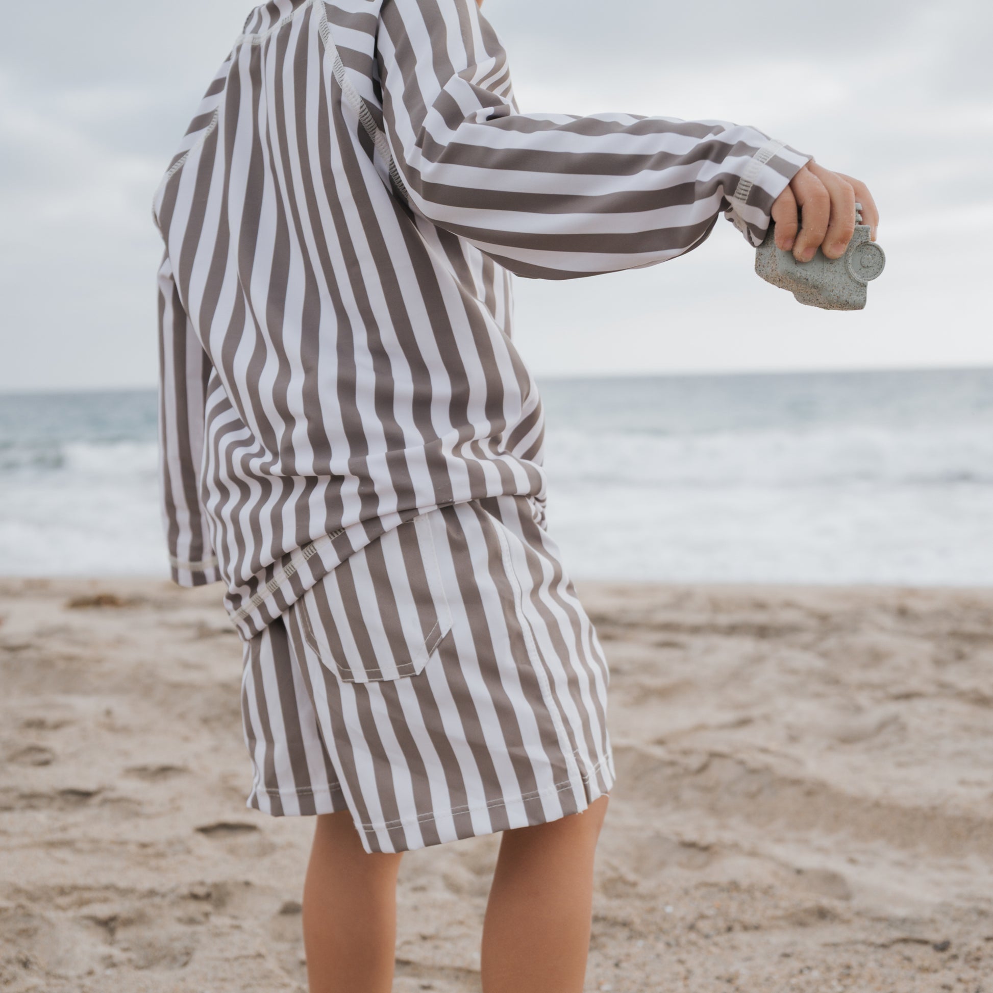 A child stands on a sandy beach facing the ocean, wearing forever french baby’s Child Board Short in Olive Stripe. The cloudy sky and sea are visible in the background as they hold a small rock.