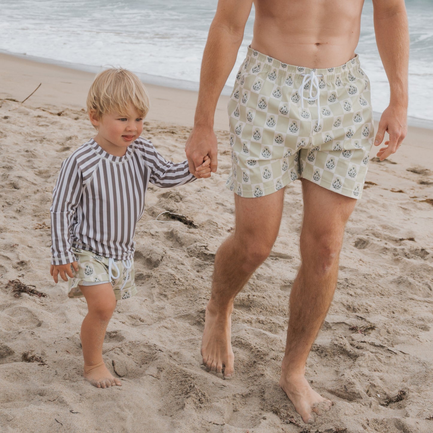A man and a boy walk barefoot along the ocean, both in beige patterned board shorts. The boy sports the forever french baby Child Board Short | Marigold Check—stylish kids activewear ideal for summer swimwear adventures.