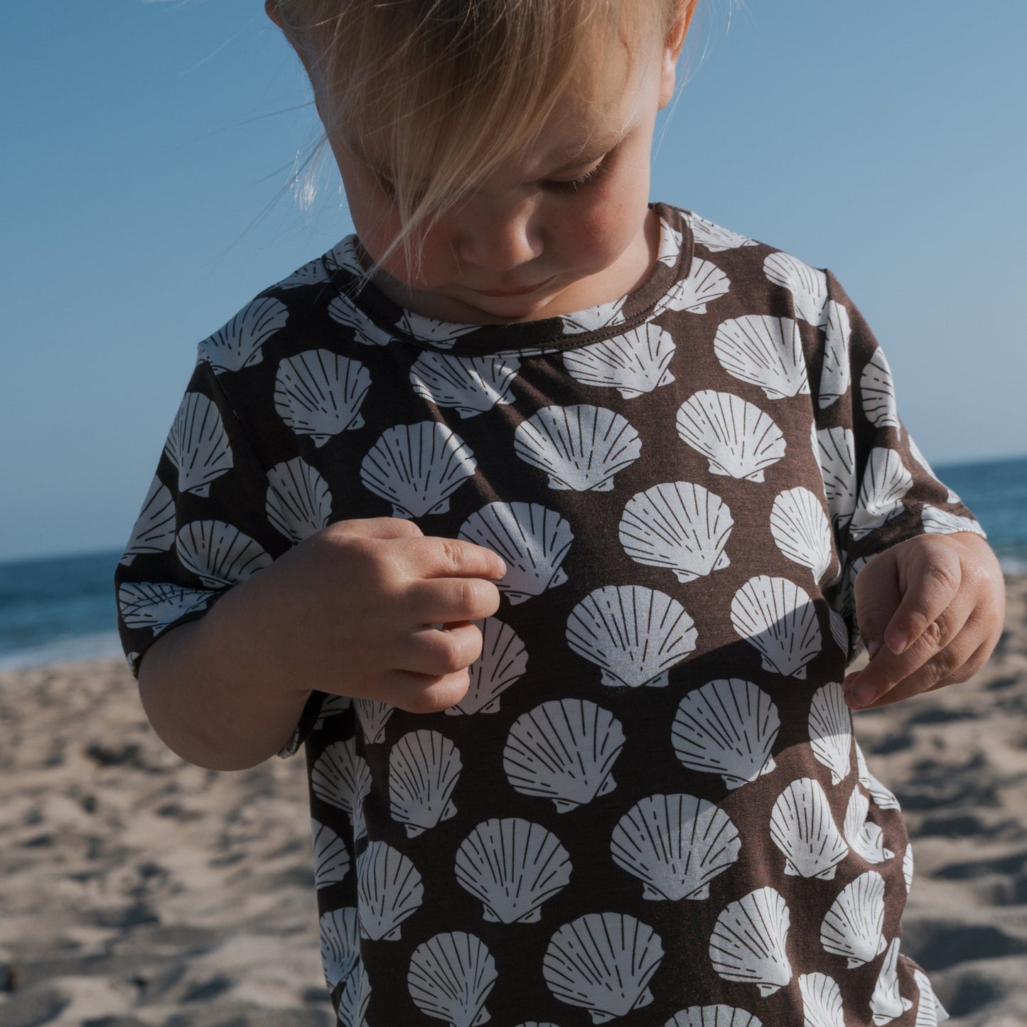 A young child in the forever french baby Bamboo Short Set | Seashells touches their brown shirt with white seashell patterns, standing on a sandy beach with the ocean and blue sky in the background.