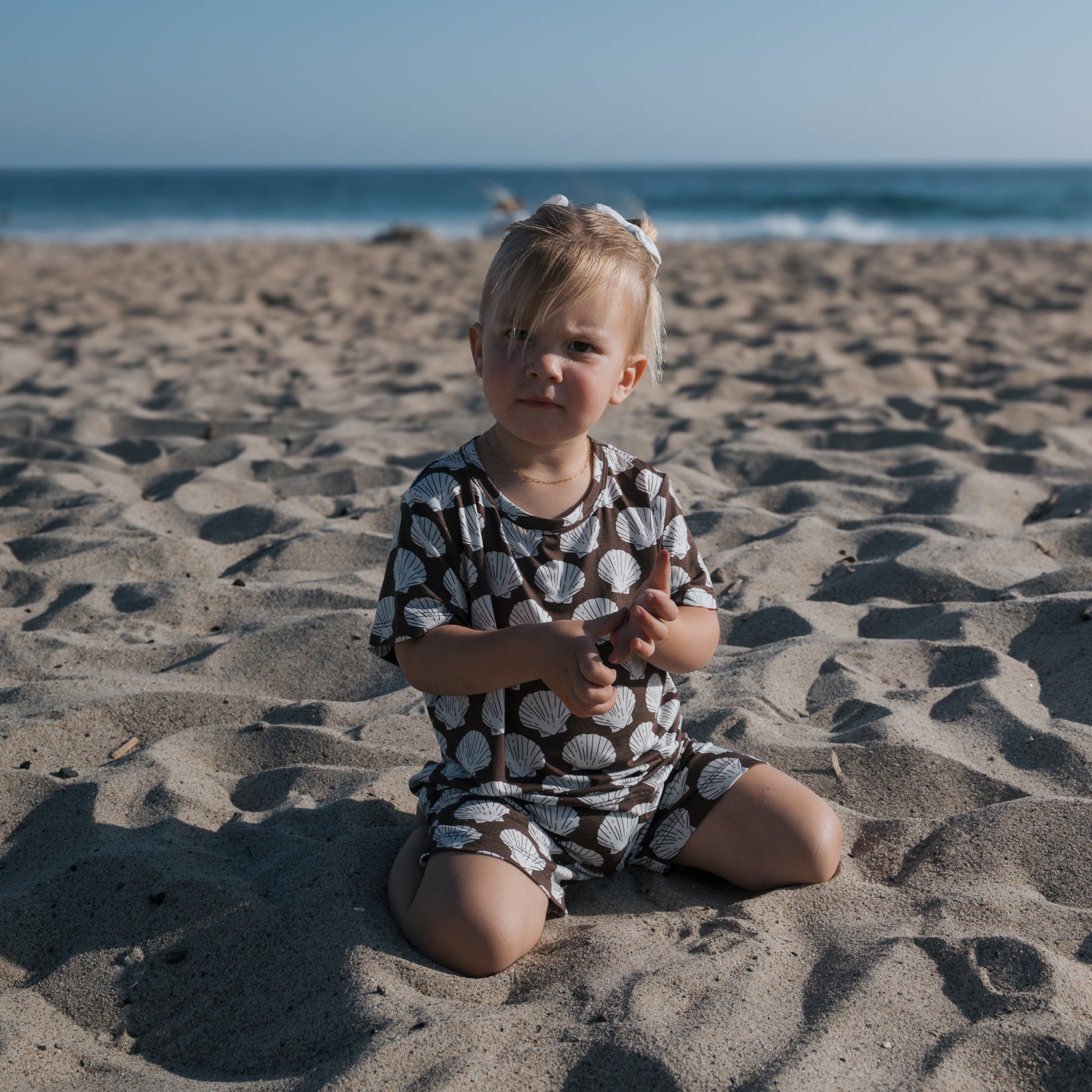 A young child wearing the forever french baby Bamboo Short Set | Seashells plays in the sand on a beach, with ocean waves and a blue sky in the background.