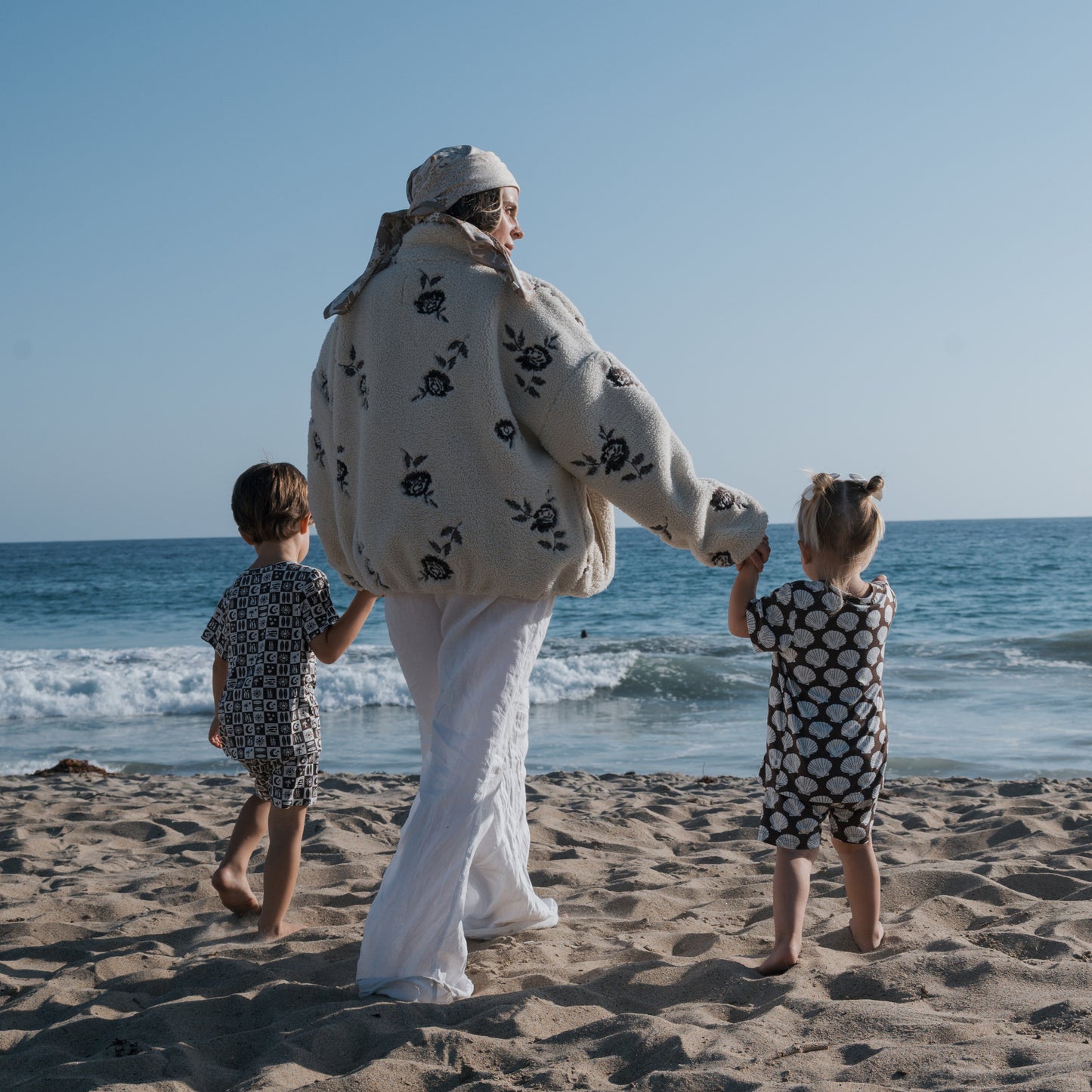 A woman in a floral jacket and headscarf walks hand-in-hand with two children on a sandy beach, both kids wearing the forever french baby Bamboo Short Set | Seashells, with gentle waves and a clear blue sky in the background.