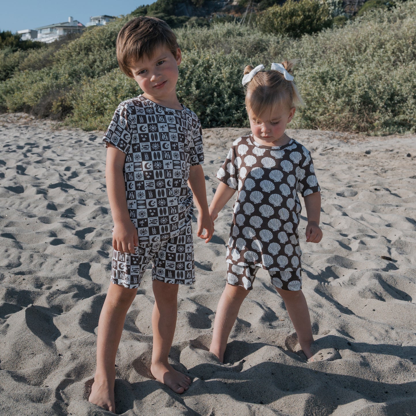 Two young children stand barefoot on a sandy beach in matching “Bamboo Short Set | Seashells” by forever french baby, featuring brown and white seashell patterns—stylish, hypo-allergenic sleepwear perfect for little ones.