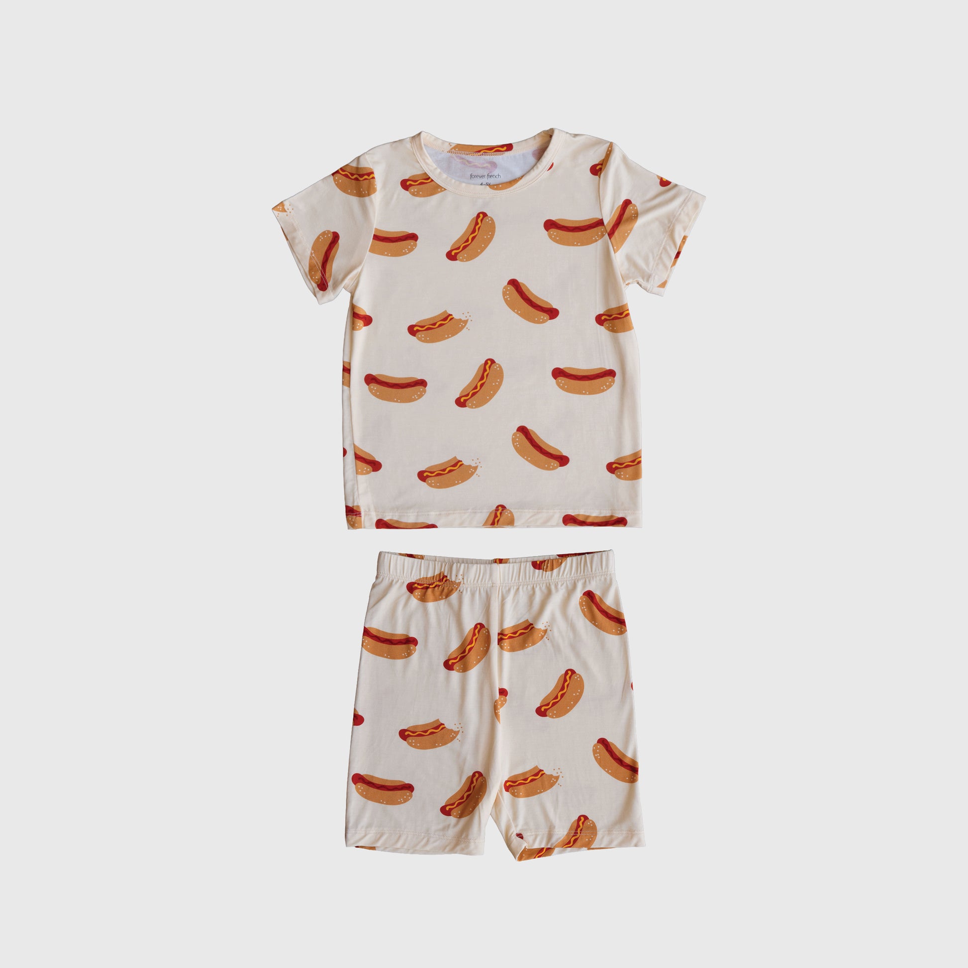 The Bamboo Short Set | Hot Dogs by forever french baby features a cream short-sleeve shirt and shorts with an all-over hot dog print, made from breathable sleepwear fabric and shown on a plain white background.