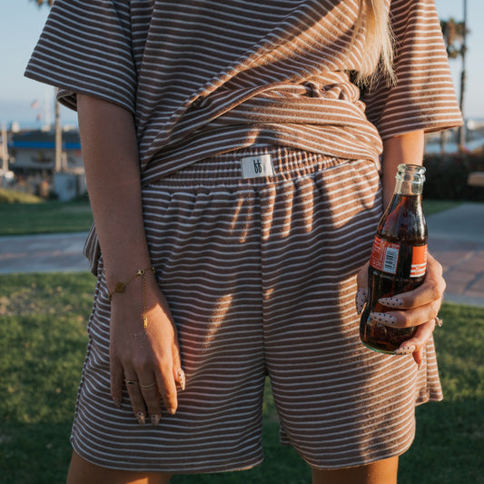 Wearing the forever french baby Women's Terry Short Set in Cola Stripe, a woman stands outdoors holding a glass bottle of soda. Sunlight highlights her painted nails, bracelet, and the comfy loungewear as shadows play across the scene.