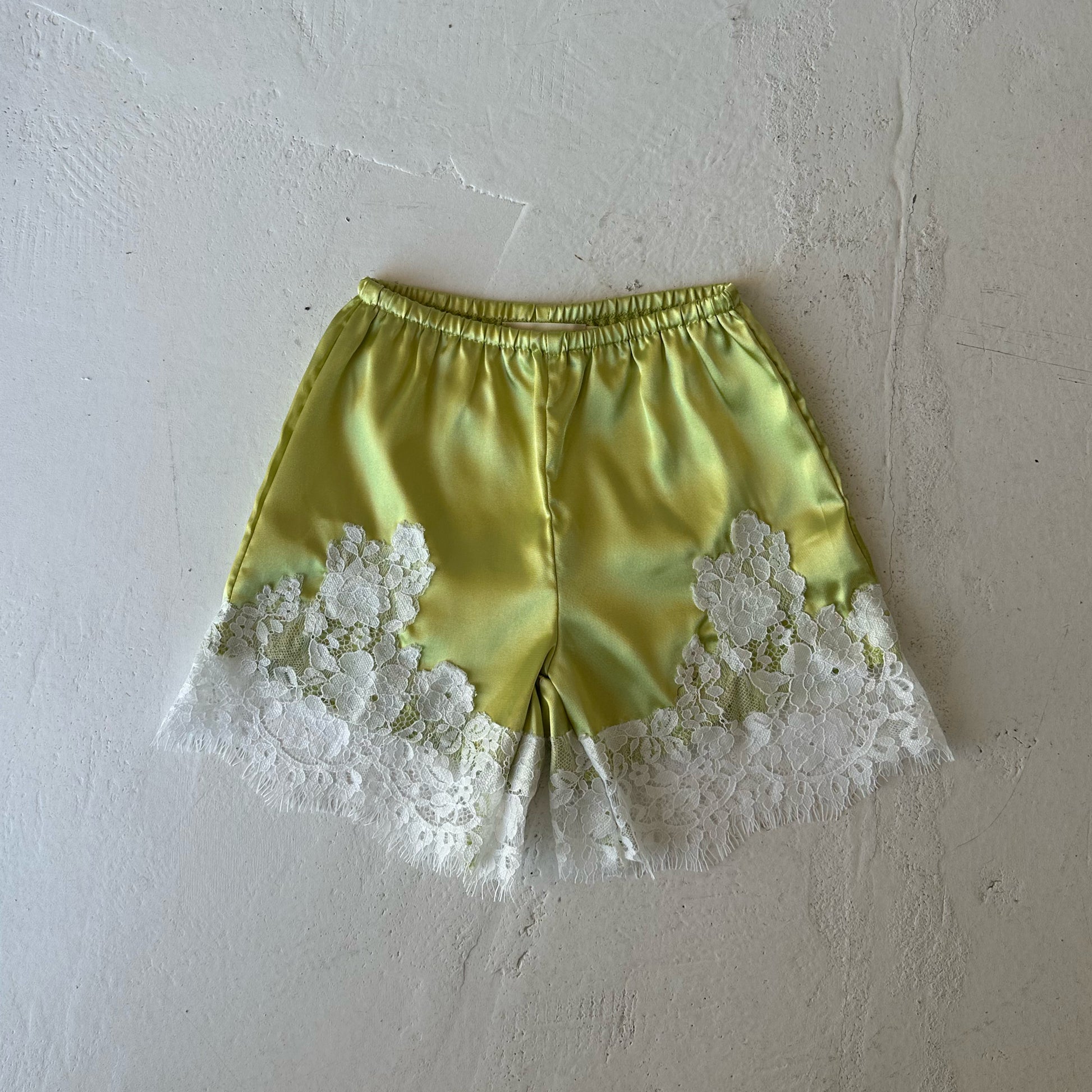 Introducing the Girl’s Satin Shorts | Prickly Pear from forever french baby—a light yellow pair of shorts with white lace trim at the hem, showcasing a delicate floral lace pattern. Designed with a lightweight satin fabric and a comfortable elastic waistband, these shorts are styled flat on a textured white surface.