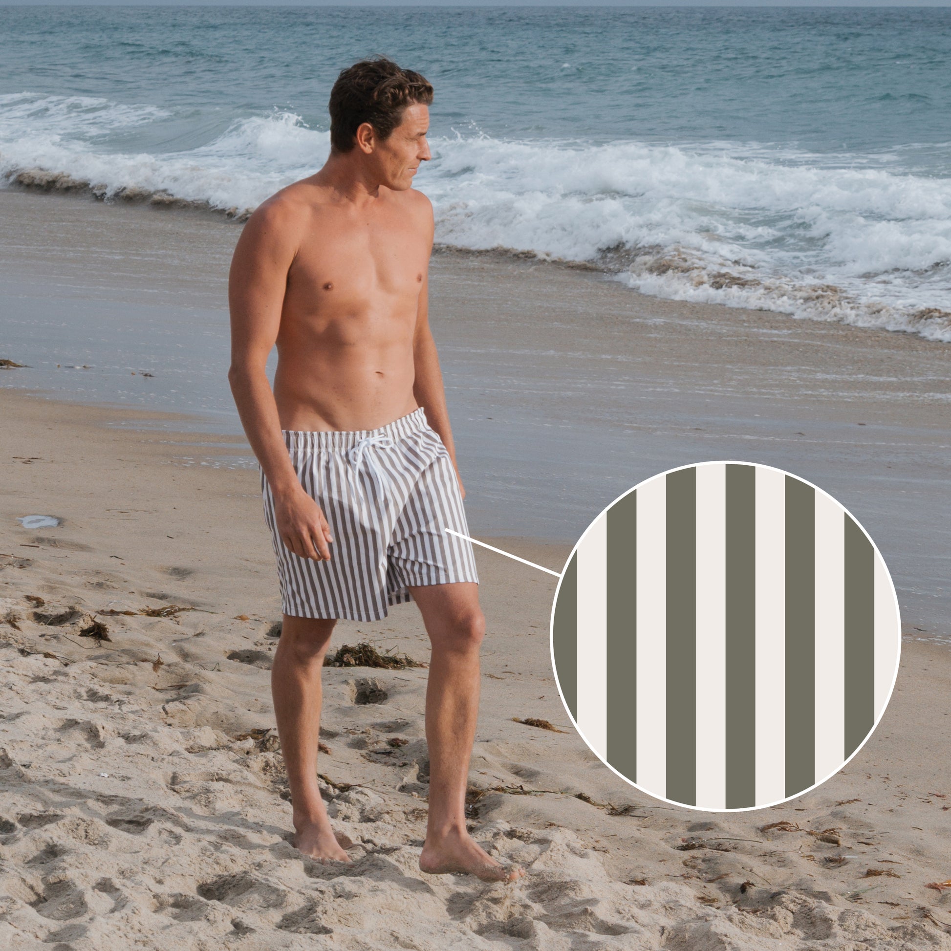 A man walks along the beach in forever french baby Men's Board Short | Olive Stripe, featuring vertical olive and white stripes. A zoomed-in circle highlights the striped pattern as waves roll in behind him.