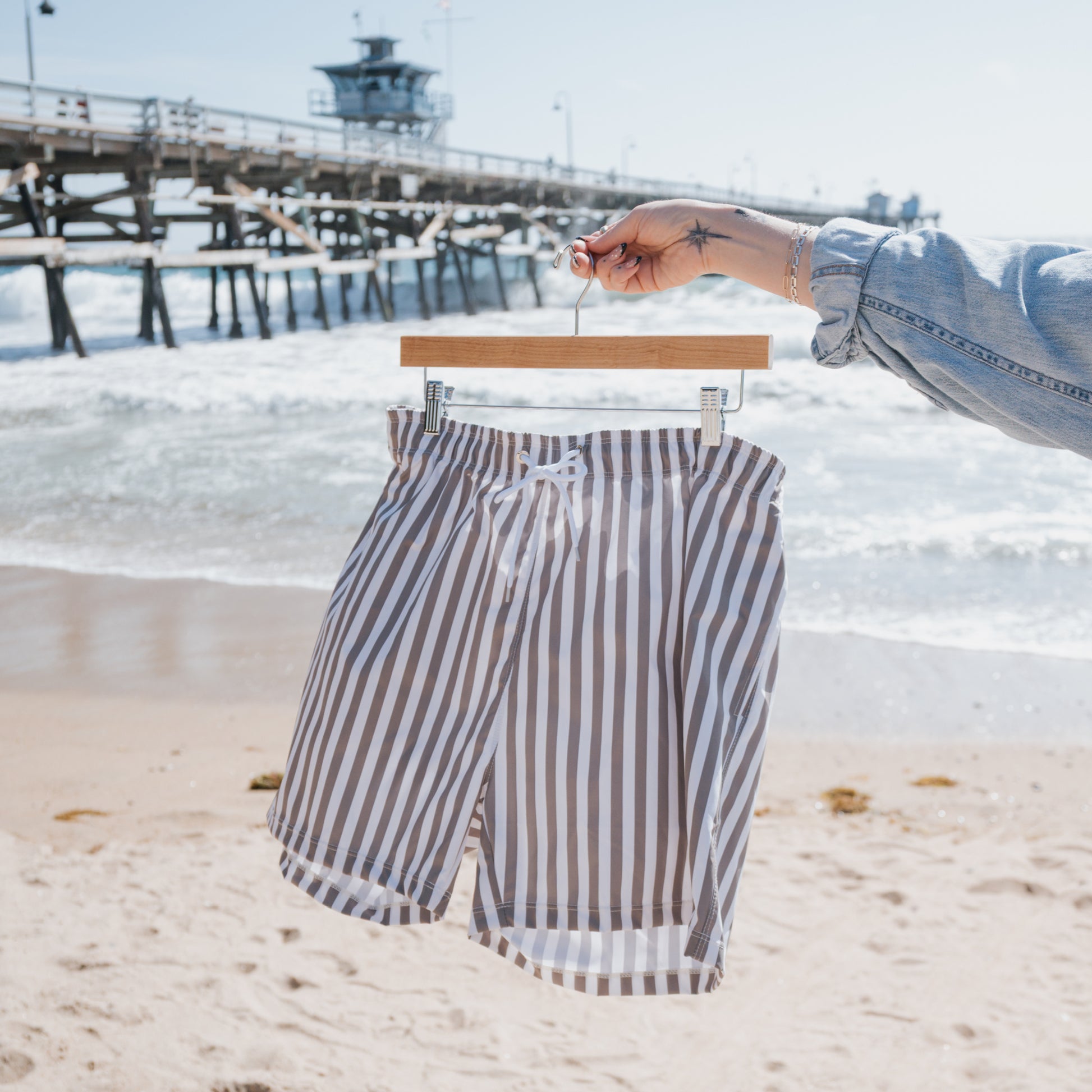 A person holds a hanger with forever french baby Men's Board Short in Olive Stripe at the sandy beach, with a wooden pier and waves in the background.