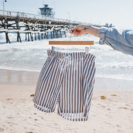 A person holds a hanger with forever french baby Men's Board Short in Olive Stripe at the sandy beach, with a wooden pier and waves in the background.