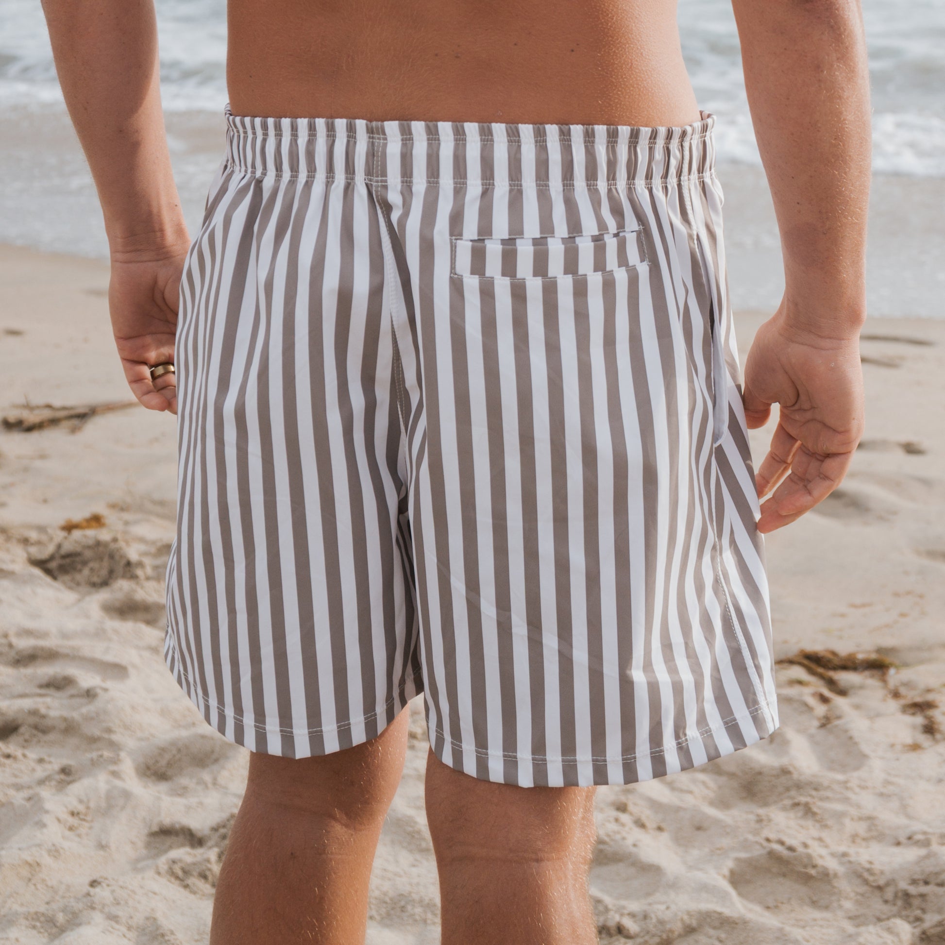 A person stands on a sandy beach facing the ocean, wearing forever french baby Men's Board Short in Olive Stripe. Only the lower back and legs are visible, set against a backdrop of sea and waves.