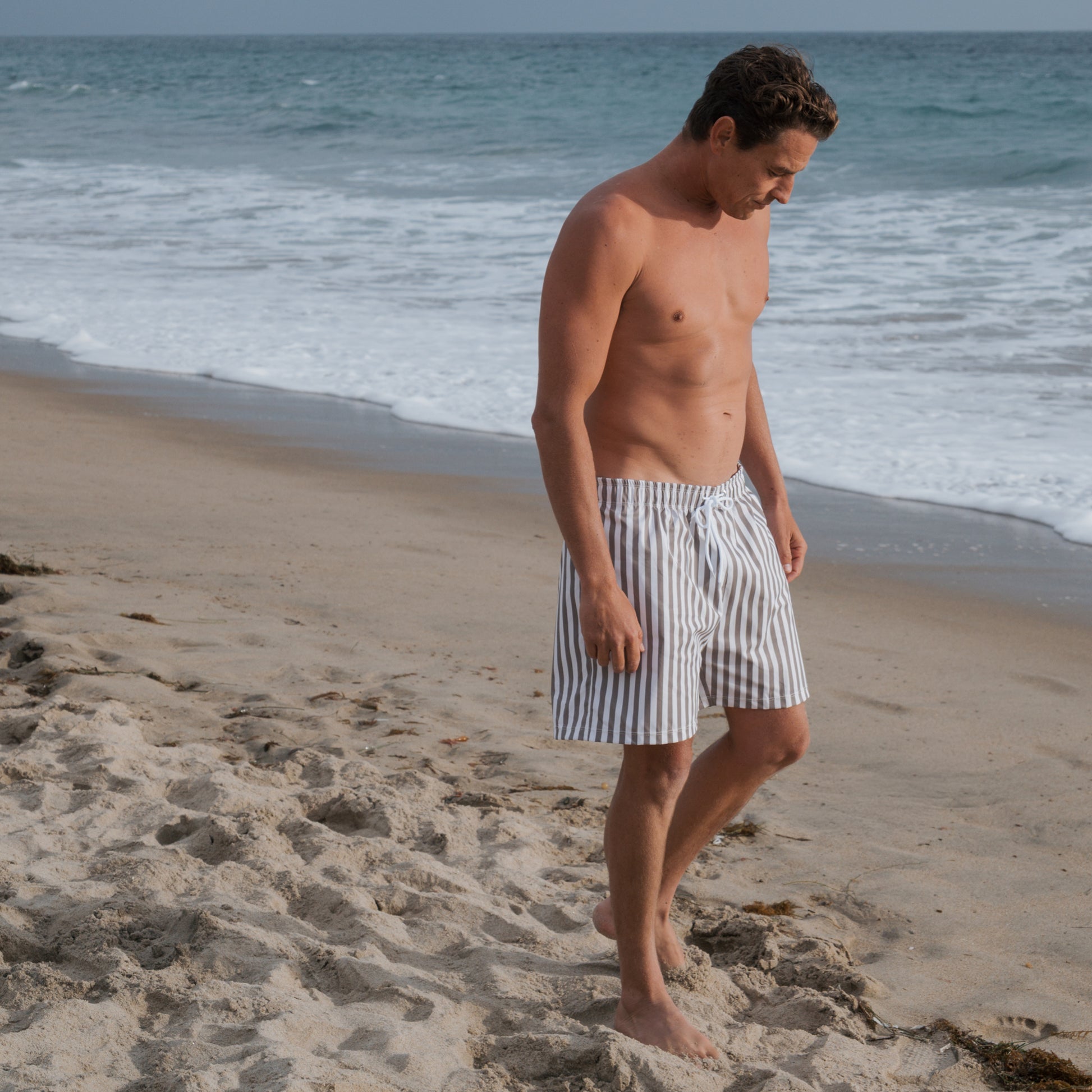 A man in forever french baby Men's Board Short | Olive Stripe walks barefoot on a sandy beach near the ocean, gazing down with waves and blue water in the background.