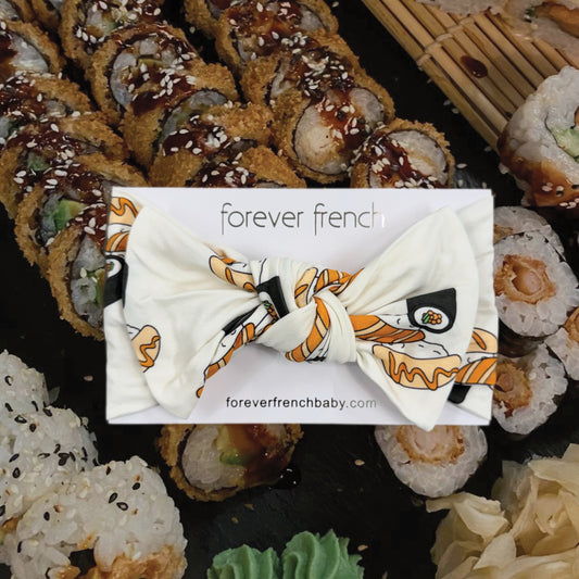 The Bamboo Head Wrap | Sushi from forever french baby is shown on a branded card, placed on a table with assorted sushi rolls and wasabi.