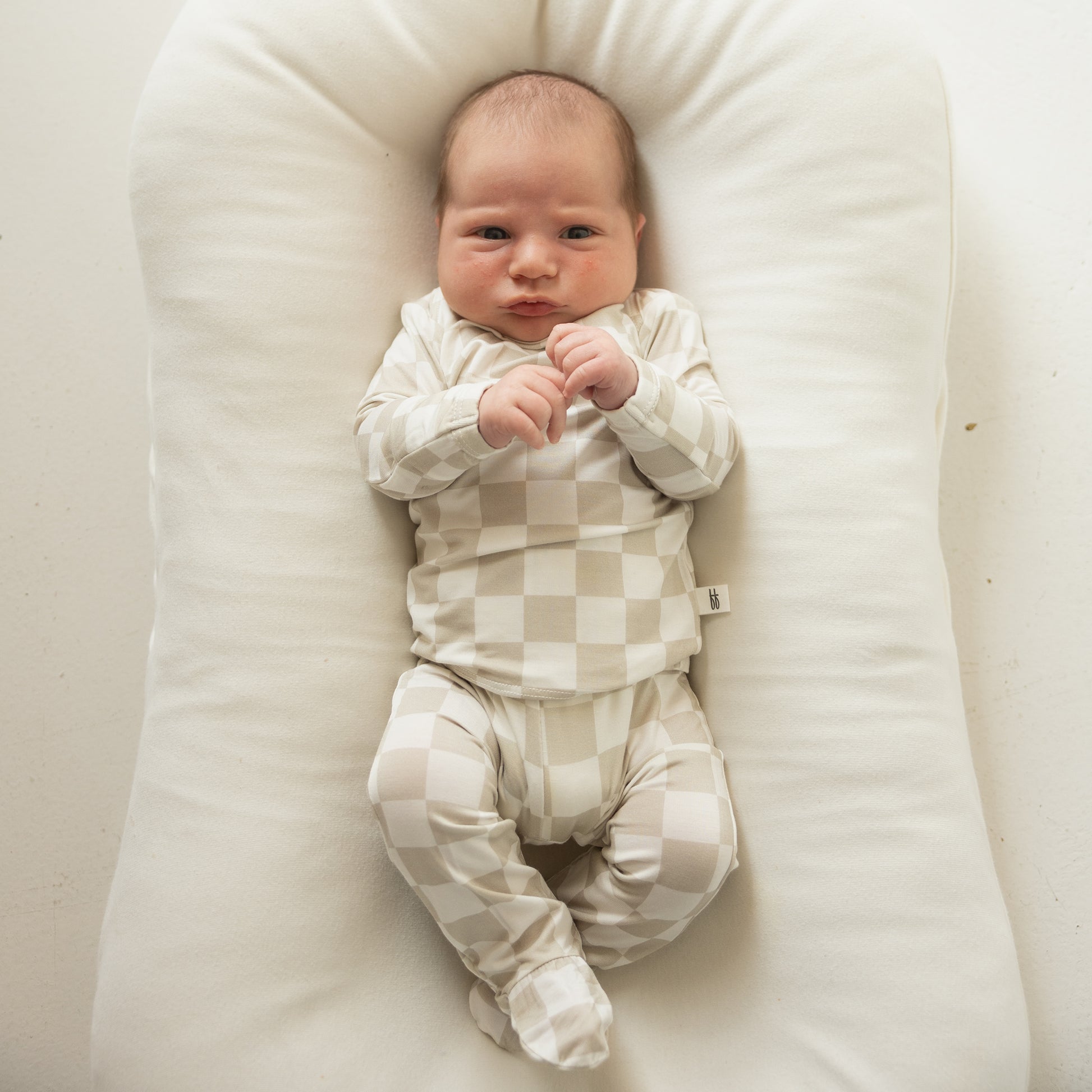 A newborn baby rests on a white cushion, wearing the Sleepy Time Set | Checkerboard—beige and white checkered long-sleeve onesie and footed pants—from forever french baby, gazing up with hands near their face.