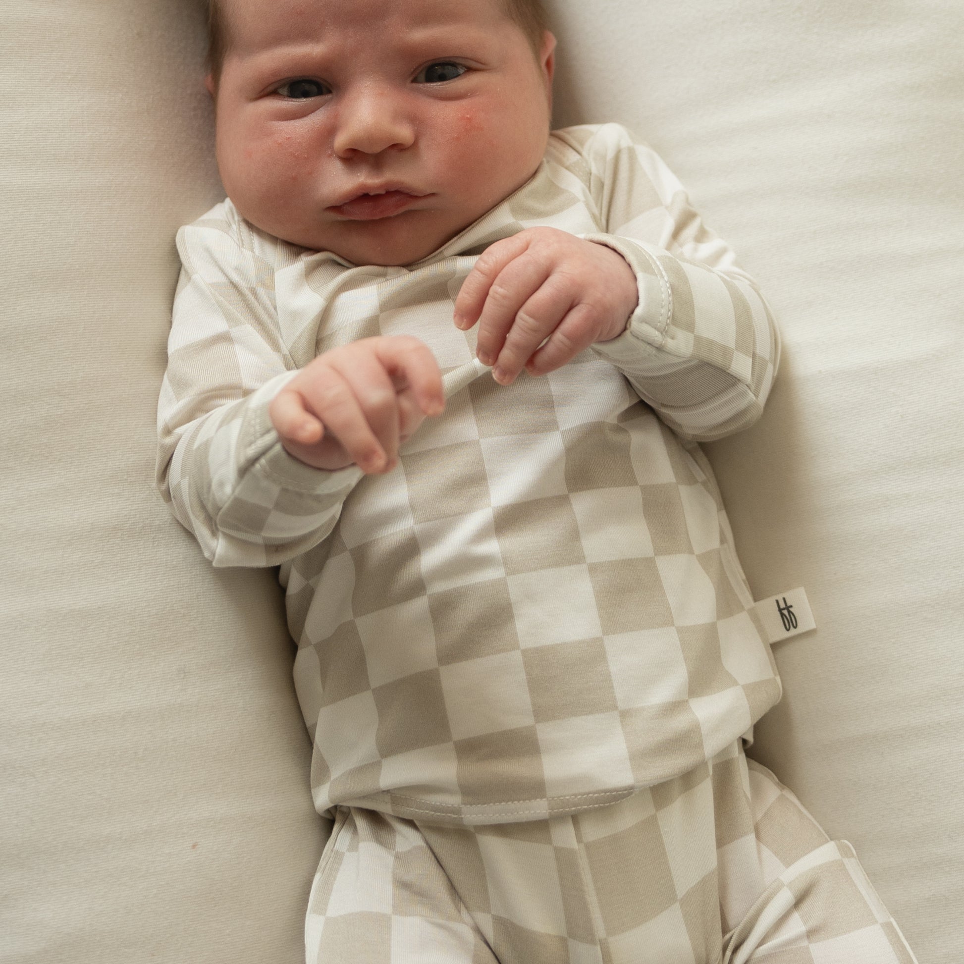 A newborn rests on a cream blanket, wearing the forever french baby Sleepy Time Set | Checkerboard in beige and white, gazing up calmly with gently clenched fists.