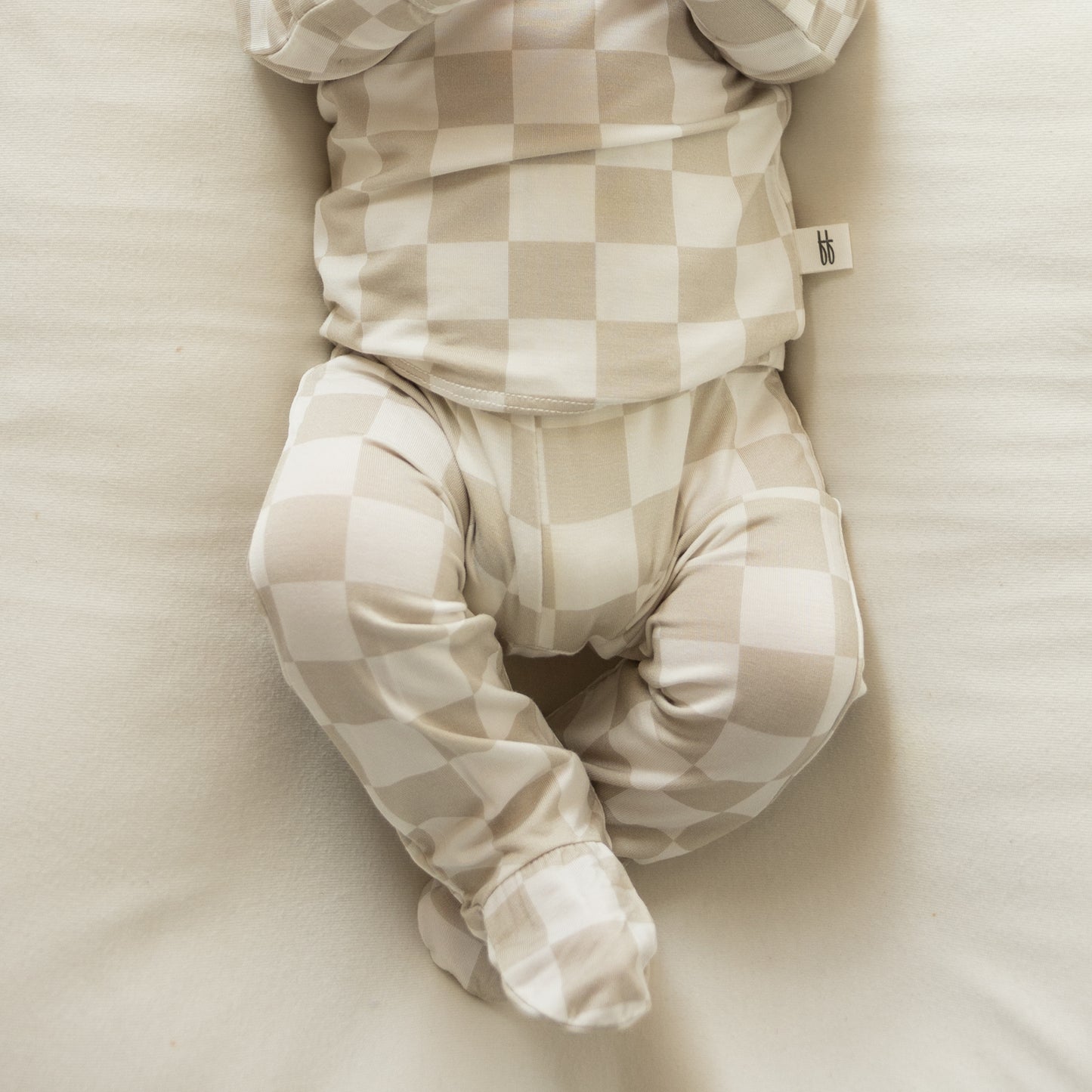 A baby lounges on a white blanket, wearing the forever french baby Sleepy Time Set | Checkerboard. Only the crossed legs and torso are visible—an ideal way to showcase this hypoallergenic outfit.