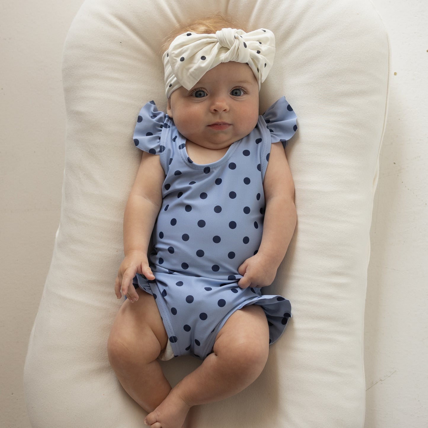 A baby in the forever French baby Girl's Sleeveless Swimsuit | Sully, featuring blue polka dots and ruffle shoulder straps, with a white headband with black dots, lies calmly on a cushioned white surface, gazing upward.