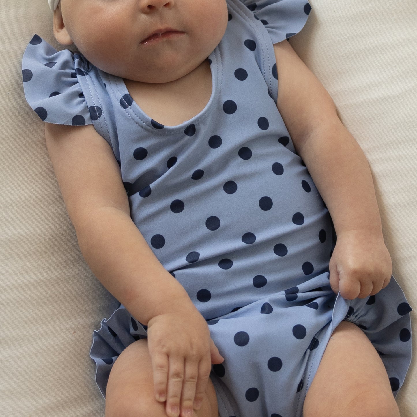 A baby wearing the forever French baby Girl's Sleeveless Swimsuit | Sully, featuring blue fabric with dark polka dots and ruffle straps, lies on a white surface while holding the swimsuit. The baby's head is partially out of frame.