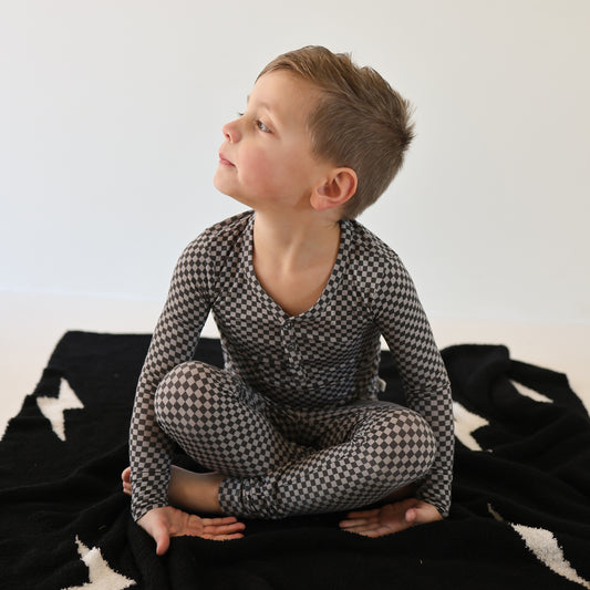 A boy wearing forever french baby Bamboo Two Piece Pajamas in Street Micro Check sits cross-legged on a black blanket with white stars, looking to his left against a plain white background.