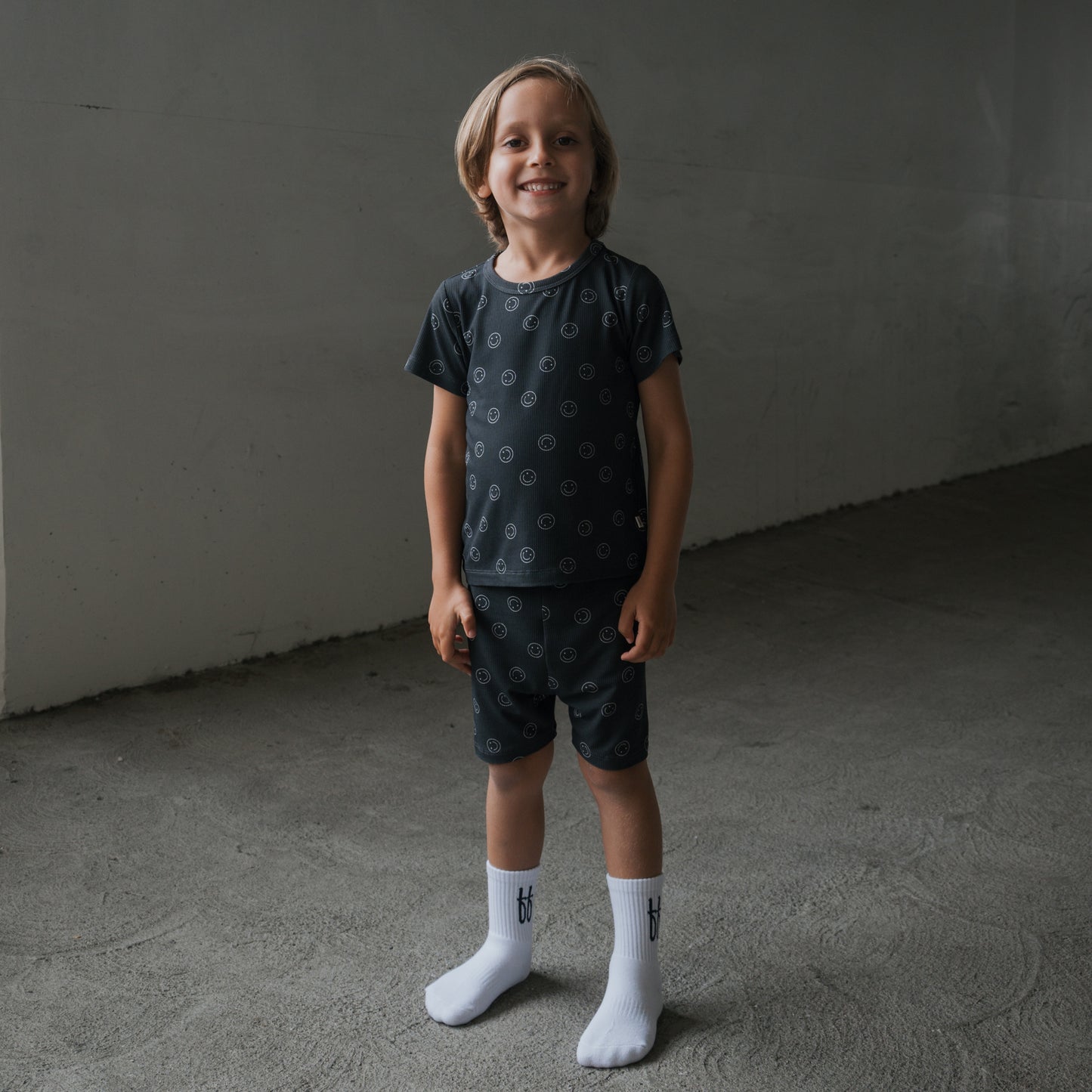 A young blonde boy smiles indoors, wearing the forever french baby Bamboo Short Set | Smiley Ribbed and white socks. He stands in a plain, gray concrete space.