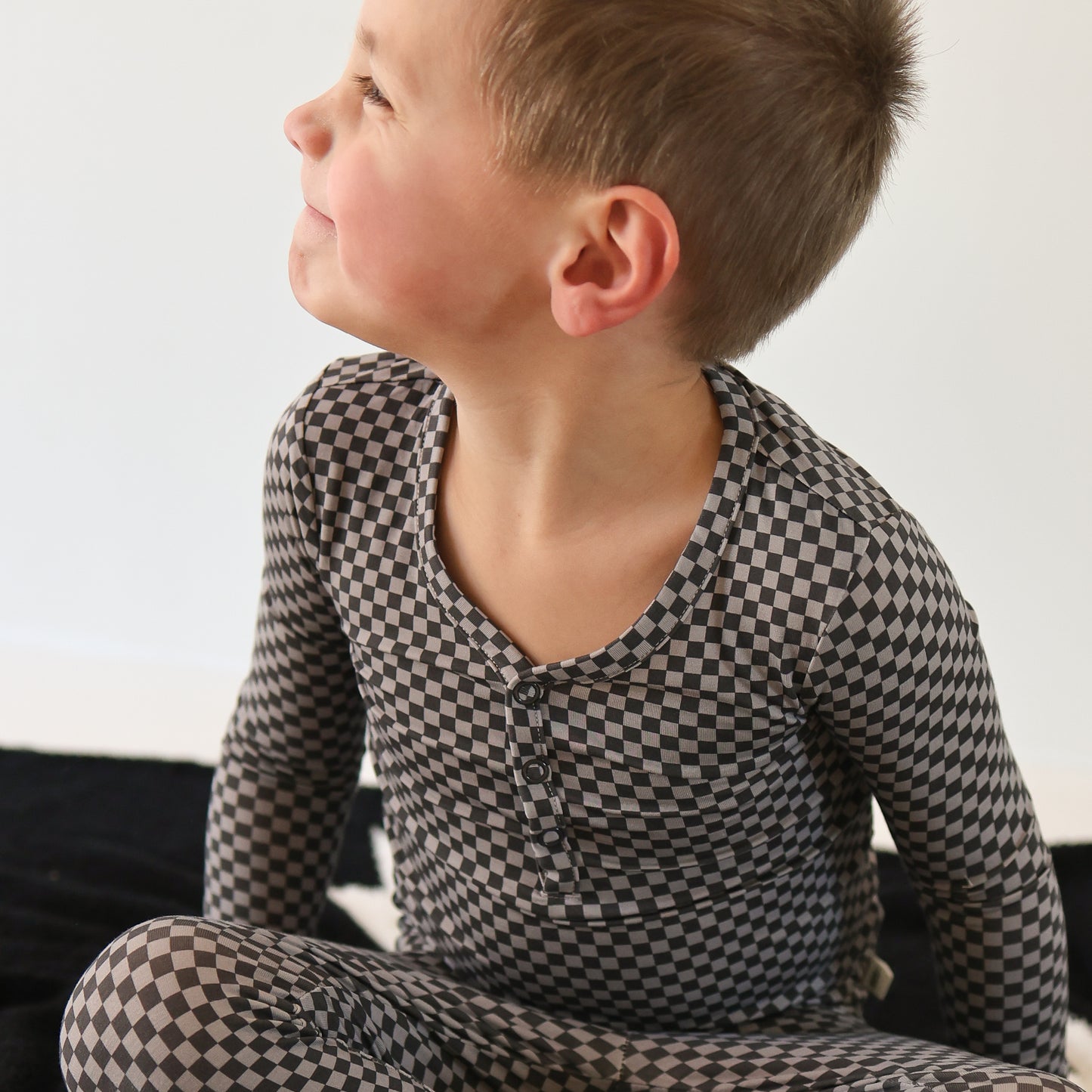 A young child sits on a dark surface, smiling and looking left while wearing forever french baby Bamboo Two Piece Pajamas in the Street Micro Check pattern. The long-sleeve, black-and-gray checkered sleepwear stands out against the plain light background.