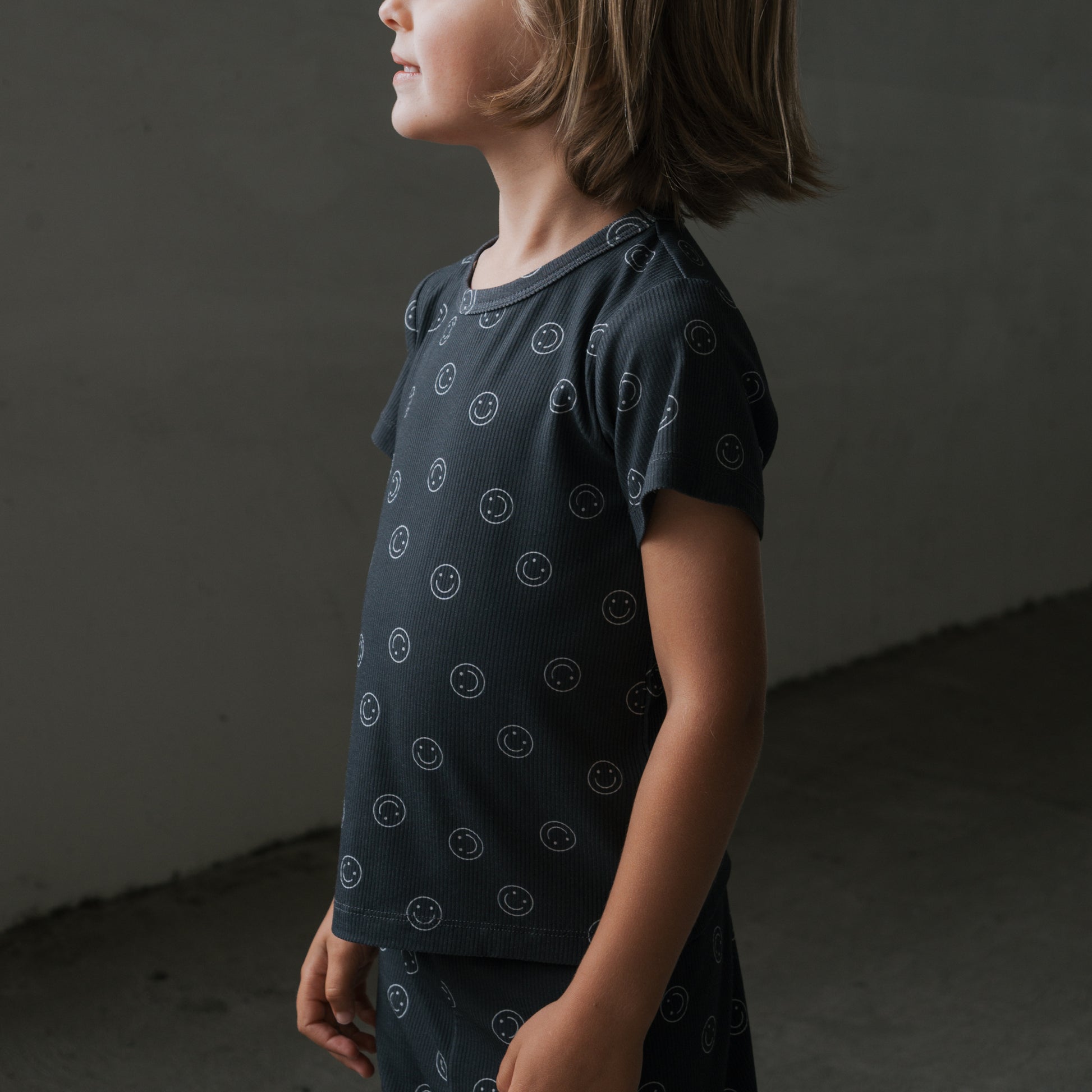 A young child with shoulder-length hair stands sideways in forever french baby's Bamboo Short Set | Smiley Ribbed—dark, hypoallergenic pajamas with a small smiley face pattern. A plain gray background highlights the comfortable, stylish sleepwear.