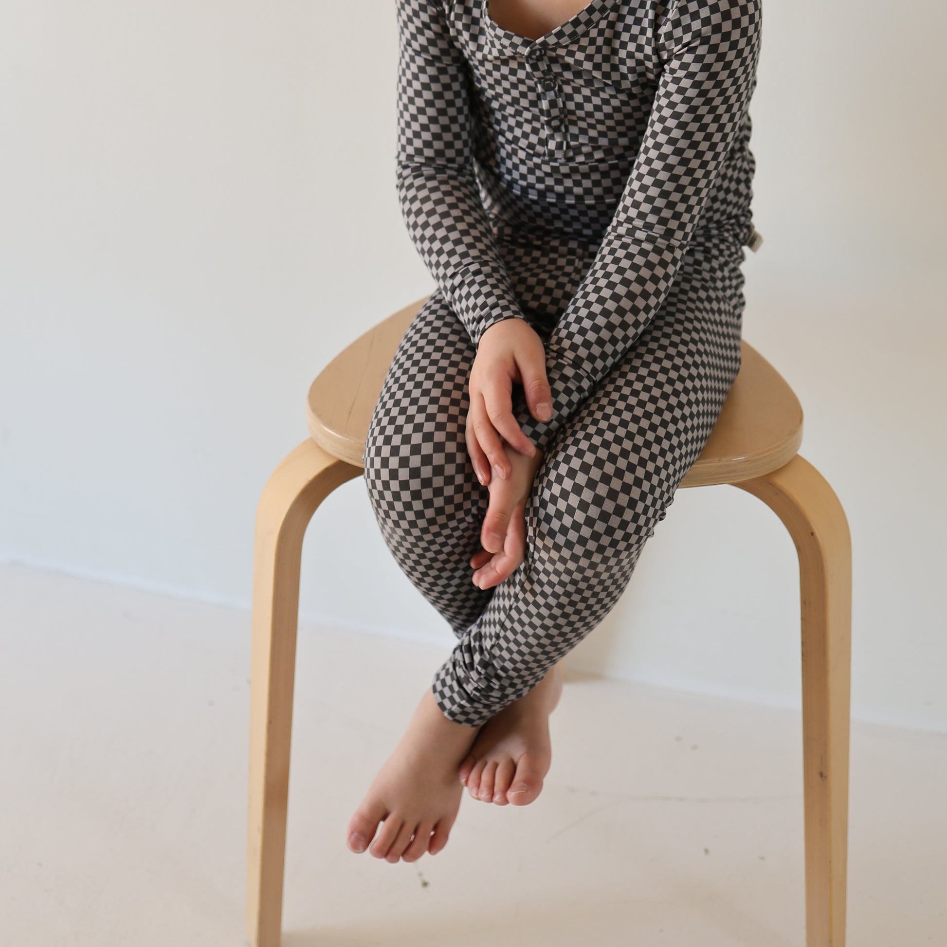A child wearing forever french baby Bamboo Two Piece Pajamas in Street Micro Check sits barefoot on a light wooden stool against a plain white background, with their face not visible.