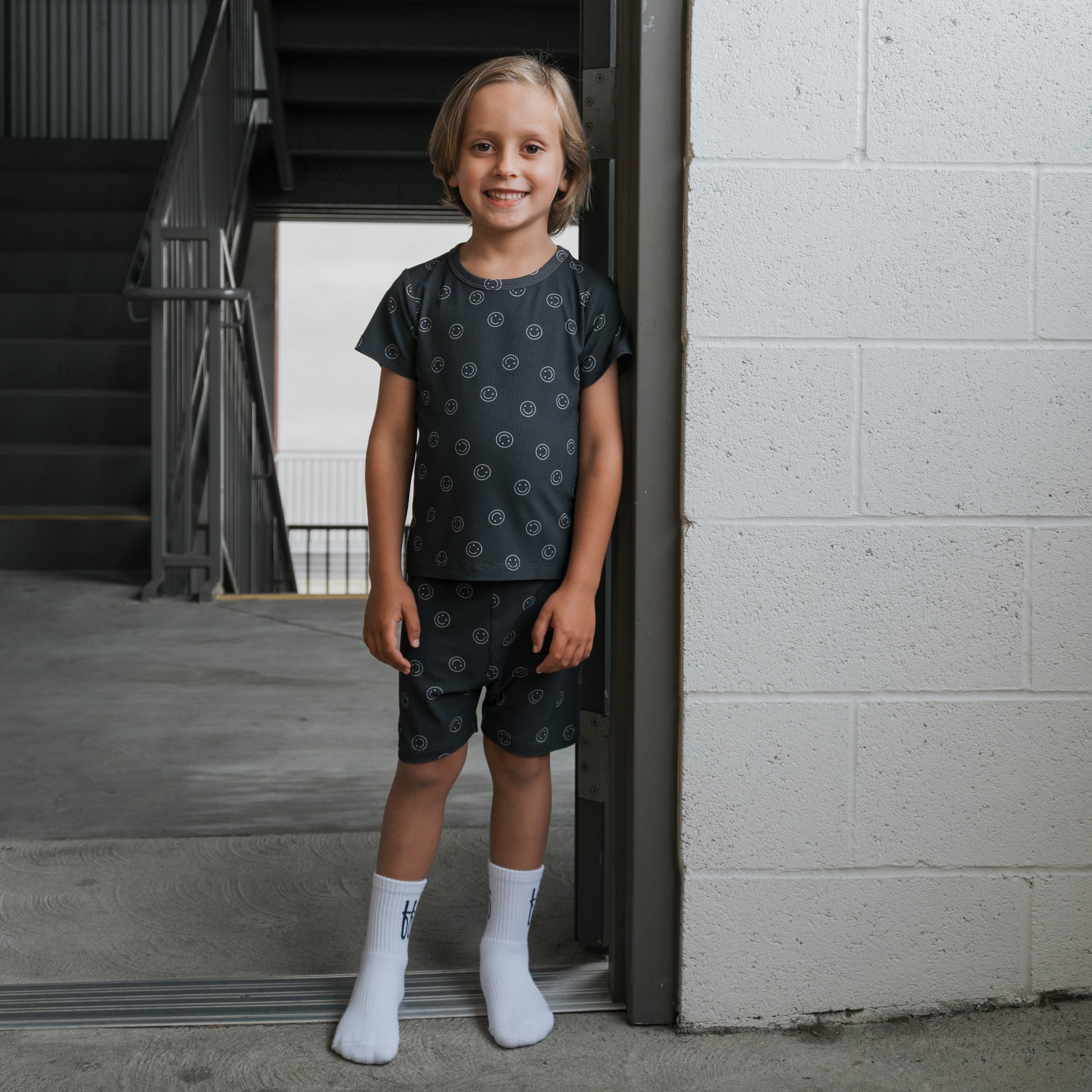 A young blonde child smiles indoors by a white brick wall, wearing forever french baby’s Bamboo Short Set | Smiley Ribbed pajamas and white socks. Stairs and a metal railing in the background highlight these cozy, hypoallergenic PJs.