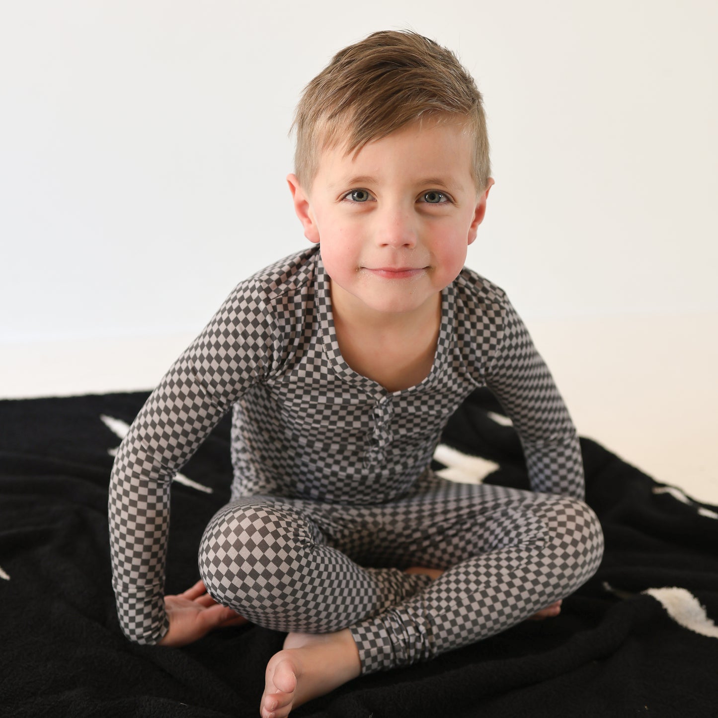 A young boy with short light brown hair sits cross-legged on a black blanket, wearing forever french baby's Bamboo Two Piece Pajamas | Street Micro Check. He smiles slightly at the camera, with a plain white background behind him.