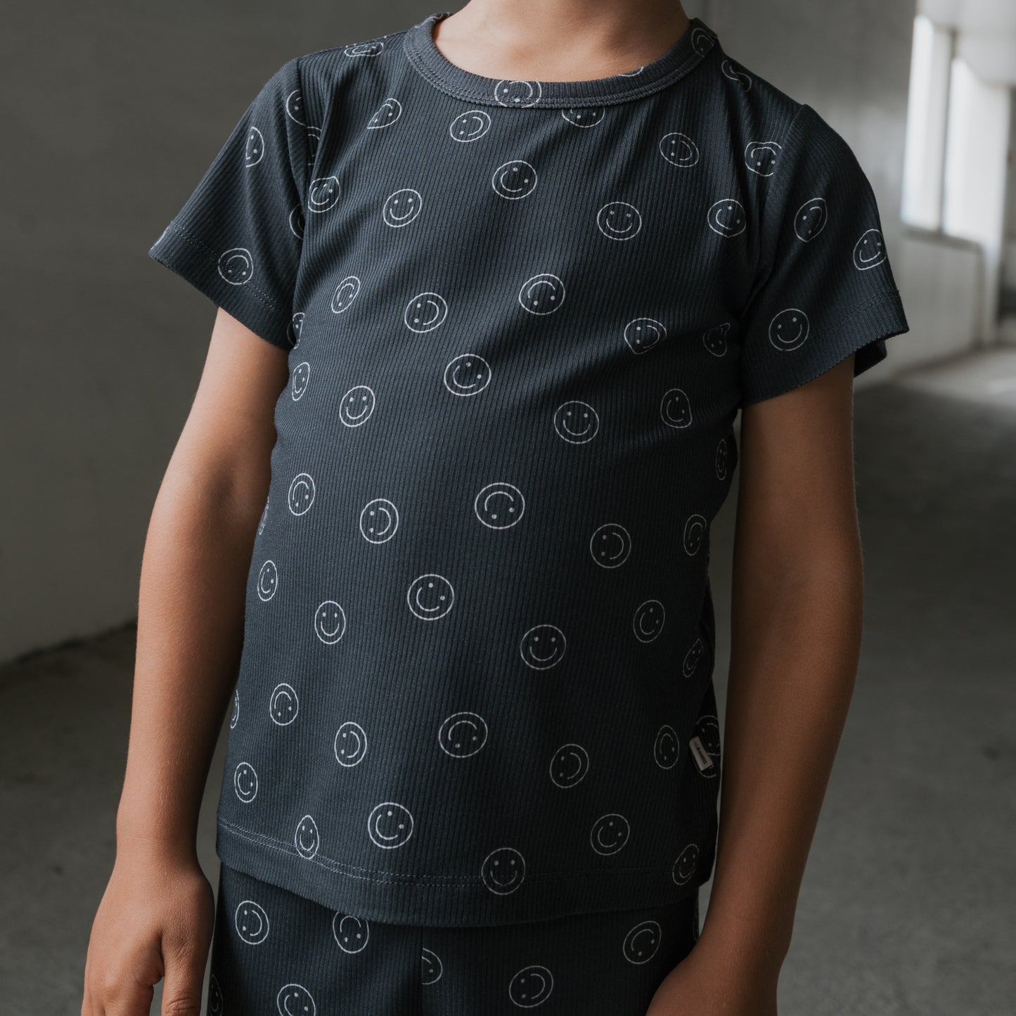 A child stands indoors in a softly lit hallway, wearing the forever french baby Bamboo Short Set | Smiley Ribbed—a dark, short-sleeved bamboo pajama top with small white smiley faces. The child's face is not visible.