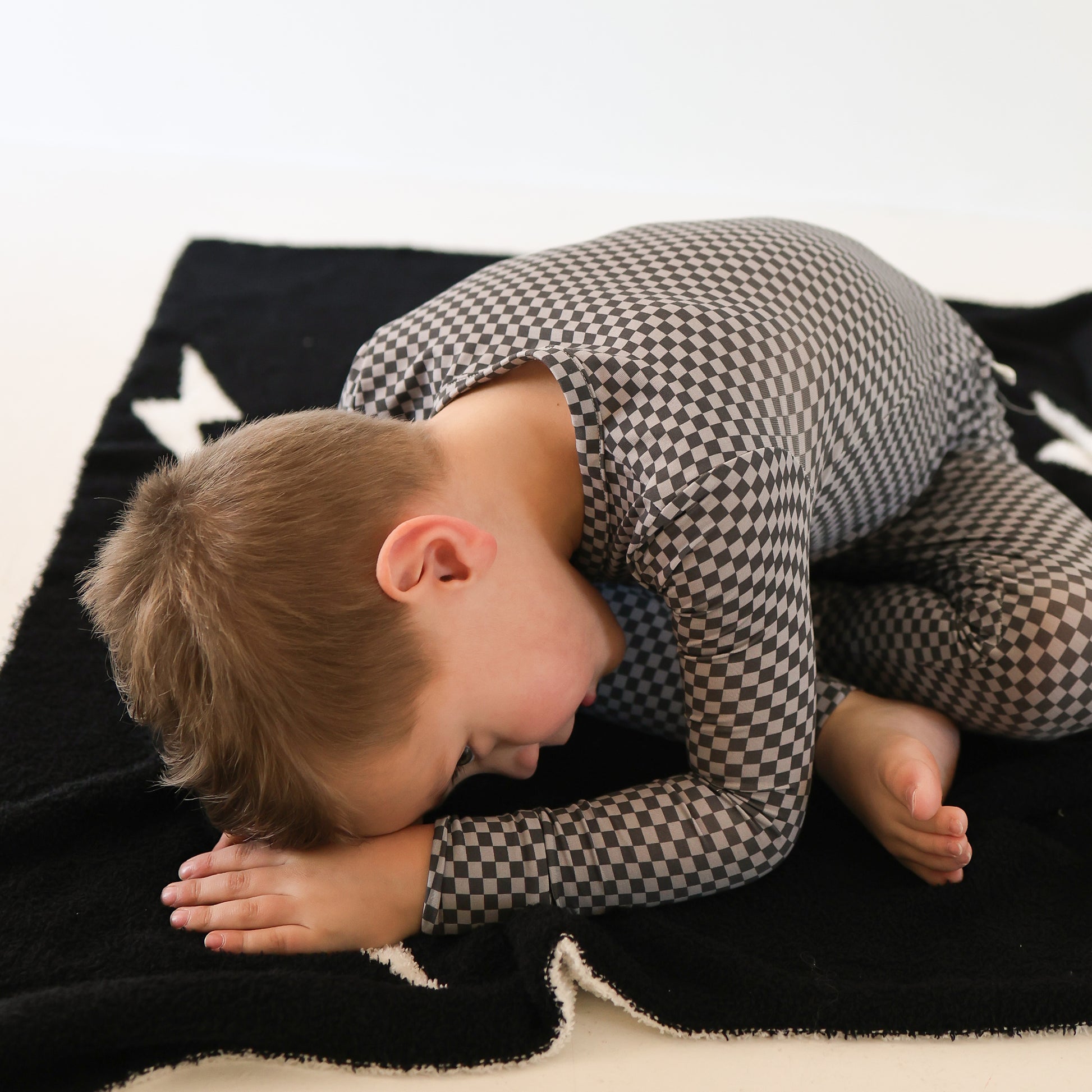 A young child in forever french baby Bamboo Two Piece Pajamas | Street Micro Check sits on a black blanket with white stars, bending forward with their forehead resting on their arms and knees.