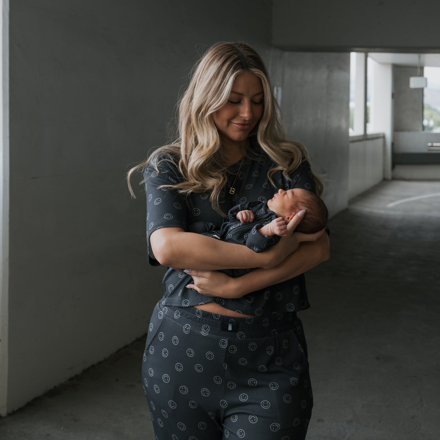 A woman with long blonde hair cradles a newborn in a dimly lit concrete parking garage. Both wear forever french baby Bamboo Zip Pajamas | Smiley Ribbed, ideal for those seeking hypo-allergenic baby clothes.