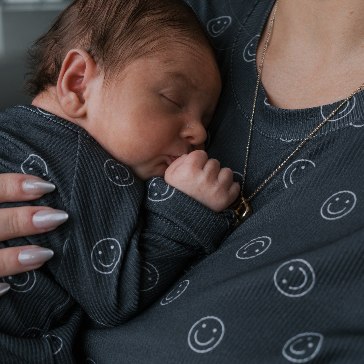A newborn sleeps in the arms of an adult, both wearing forever french baby Bamboo Zip Pajamas | Smiley Ribbed—dark gray pajamas with white smiley faces. The adult’s manicured hand supports the thumb-sucking baby.