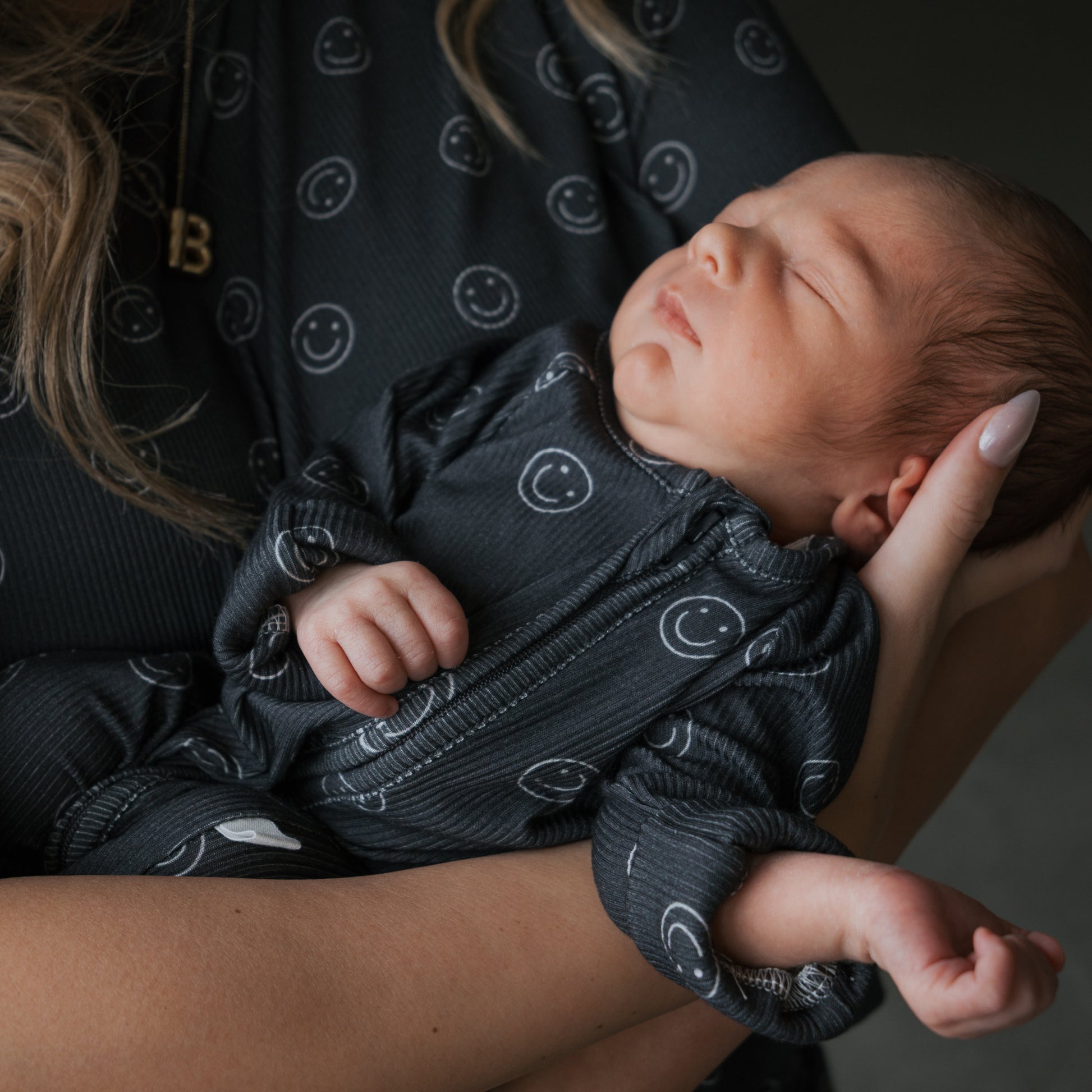 A newborn sleeps peacefully in someone's arms, wearing forever french baby's Bamboo Zip Pajamas | Smiley Ribbed. An adult supports the baby, with part of a necklace visible on their neck.