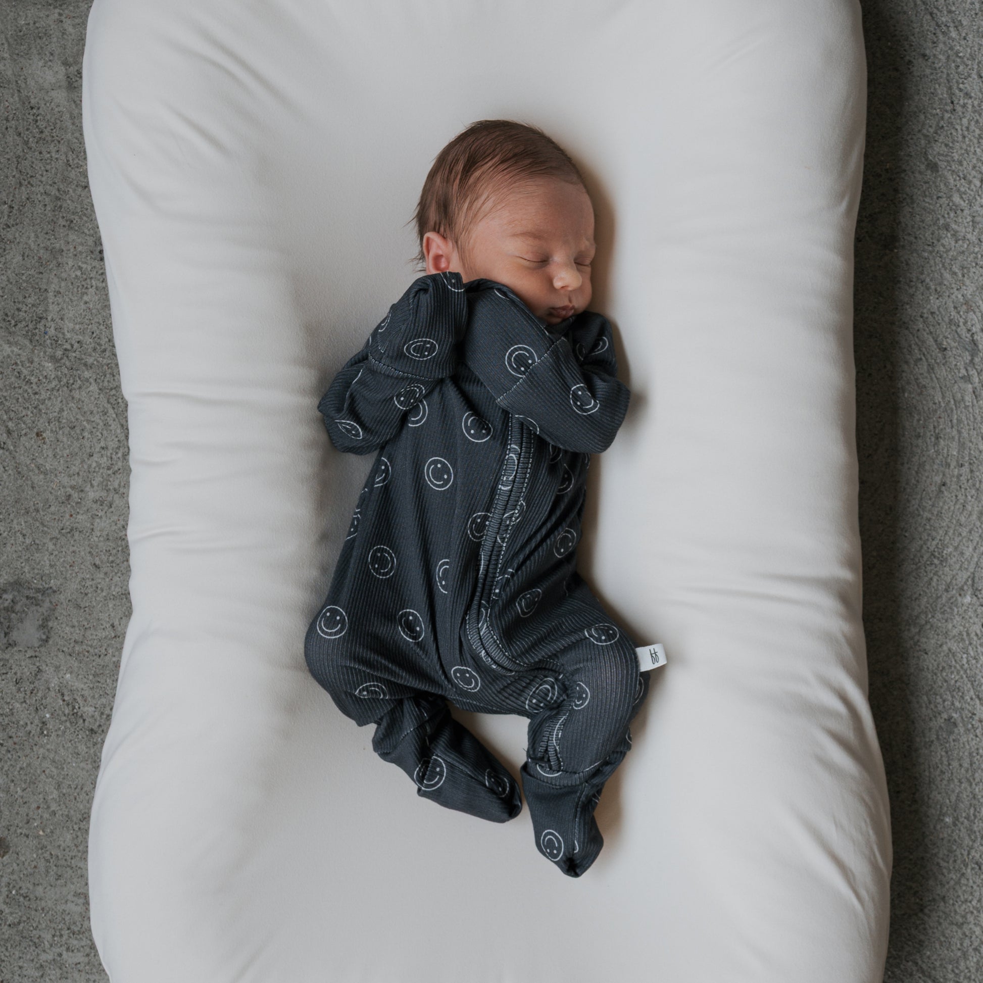 A newborn sleeps peacefully on a white cushioned pad, wearing forever french baby's Bamboo Zip Pajamas | Smiley Ribbed in dark blue with a subtle pattern, lying on their back with arms up and legs slightly bent.