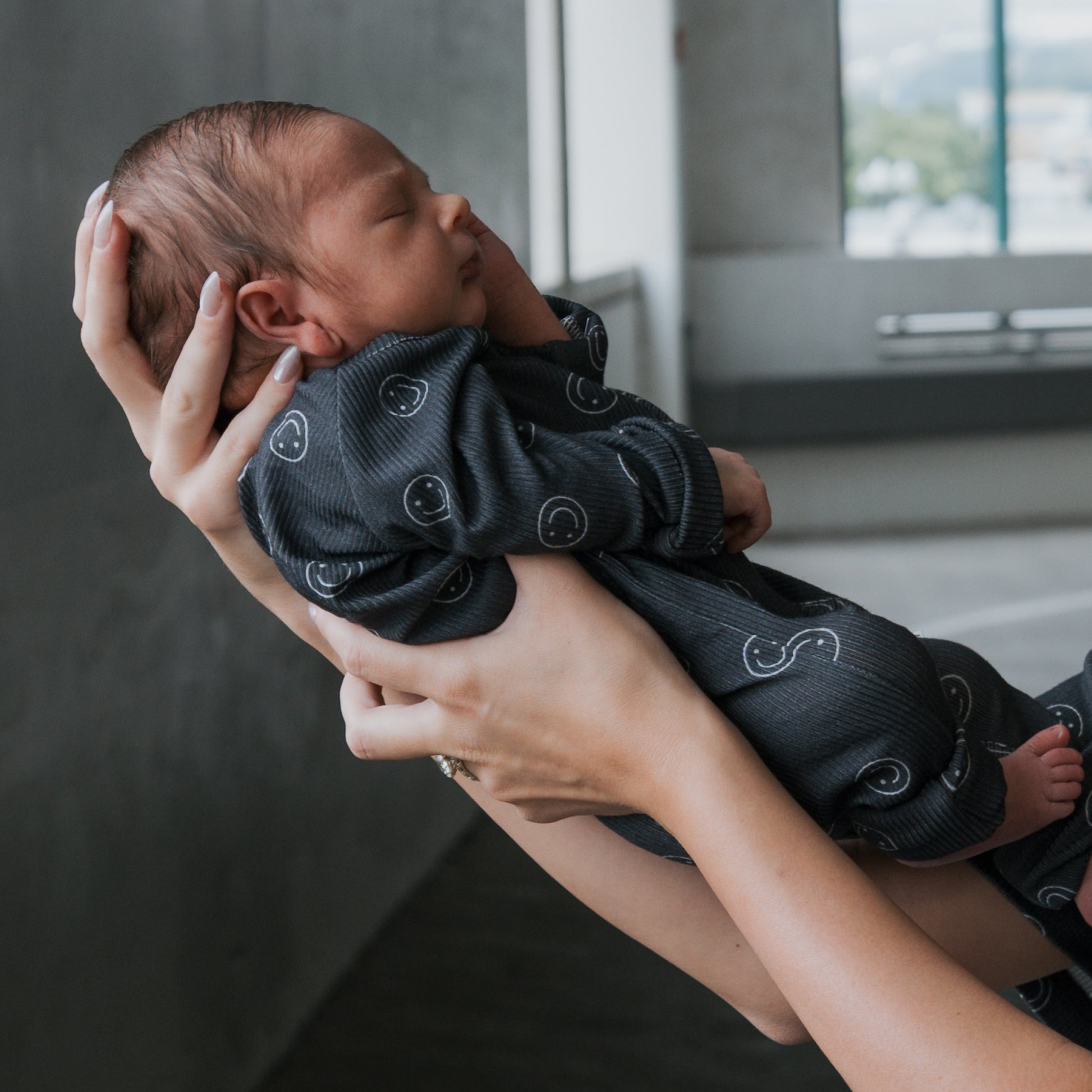 A newborn, gently cradled in adult hands, wears forever french baby's Bamboo Zip Pajamas | Smiley Ribbed, set against a softly blurred indoor background.