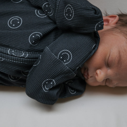 A newborn sleeps peacefully on a white surface, dressed in forever french baby Bamboo Zip Pajamas | Smiley Ribbed—dark, ribbed bamboo pajamas with white smiley face patterns.
