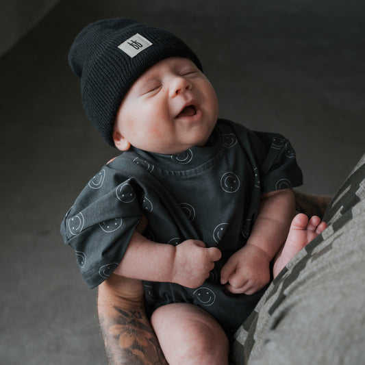 A smiling baby in a black beanie and the forever french baby Short Sleeve Romper | Smiley, covered in smiley faces, sits contentedly in tattooed arms with eyes closed.