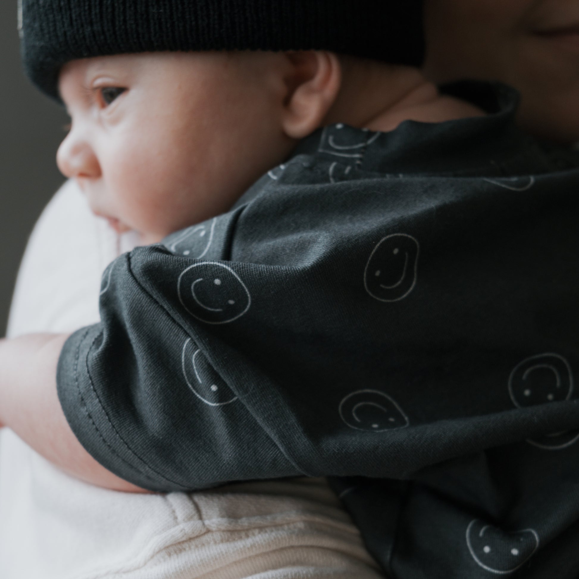 A baby in forever french baby's Short Sleeve Romper | Smiley and a black beanie rests on an adult’s shoulder, gazing to the side as gentle natural light highlights their face.