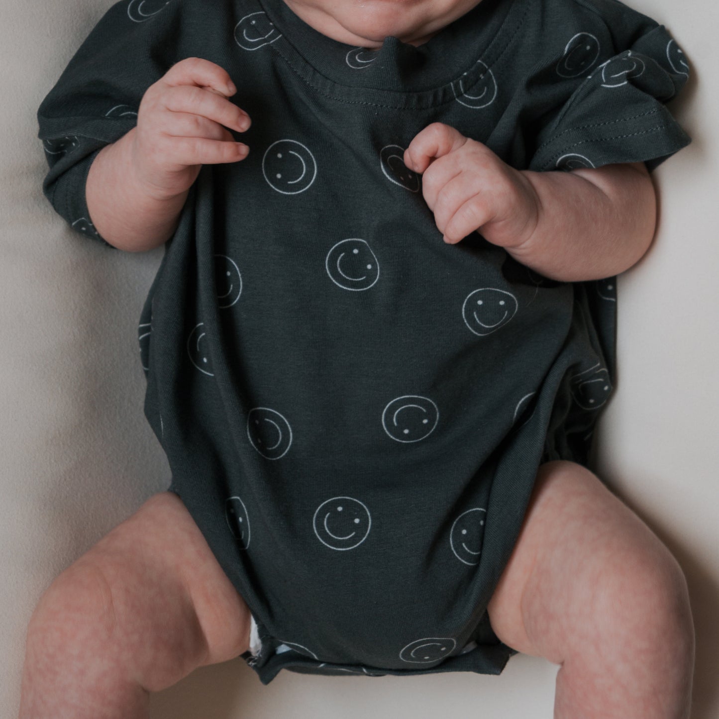 A baby in the forever french baby Short Sleeve Romper | Smiley, featuring a dark cotton fabric with white smiley face patterns, lies on a light surface with arms slightly raised and relaxed legs. The baby's face is partially out of frame.