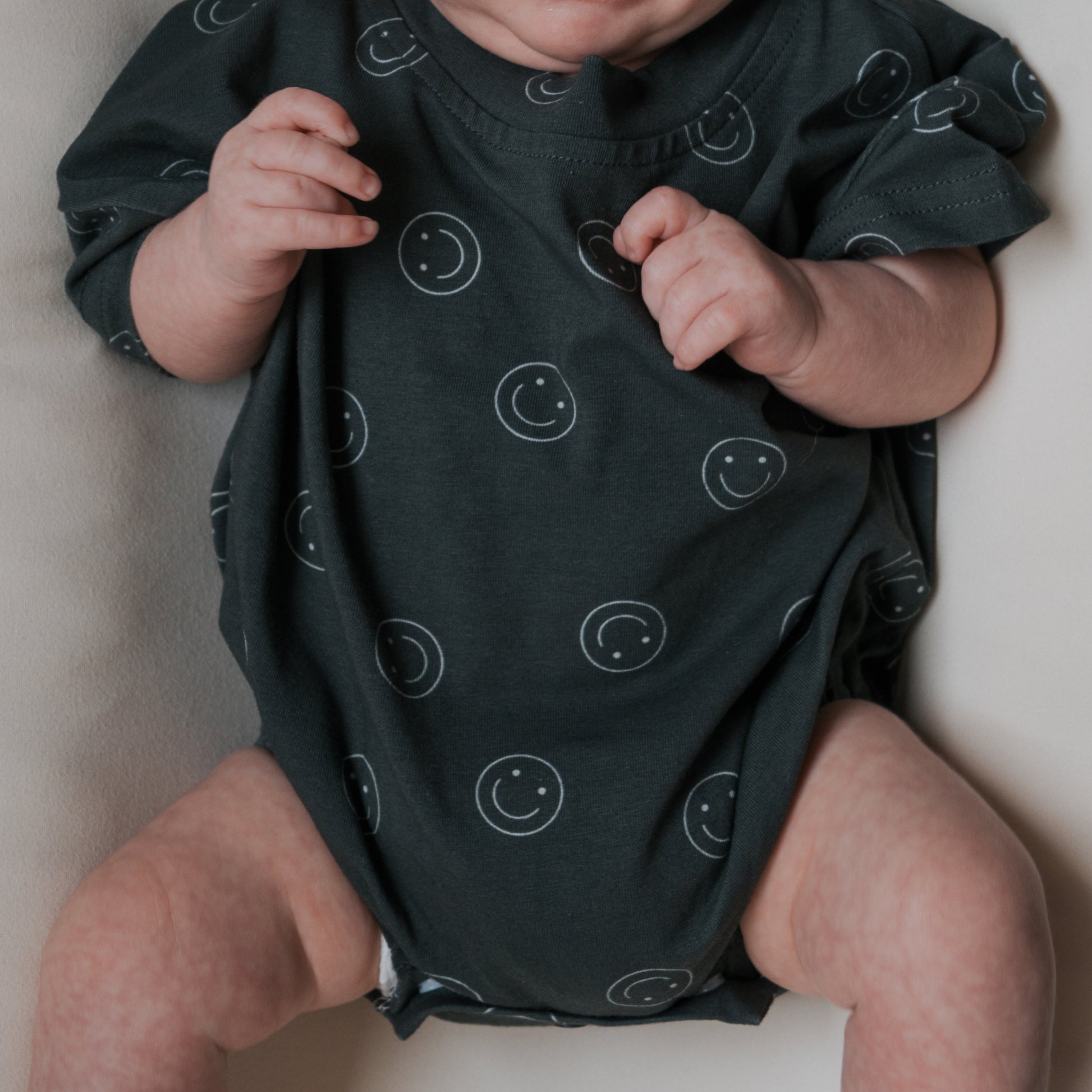 A baby in the forever french baby Short Sleeve Romper | Smiley, featuring a dark cotton fabric with white smiley face patterns, lies on a light surface with arms slightly raised and relaxed legs. The baby's face is partially out of frame.