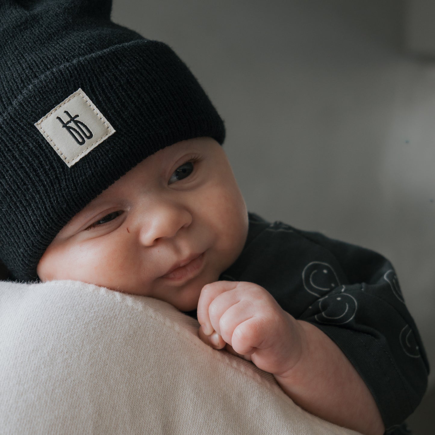 A baby in the forever french baby Short Sleeve Romper | Smiley and a black knit hat with a white logo label rests contentedly on someone's shoulder, hand gently clasped, in a warmly lit, inviting setting.