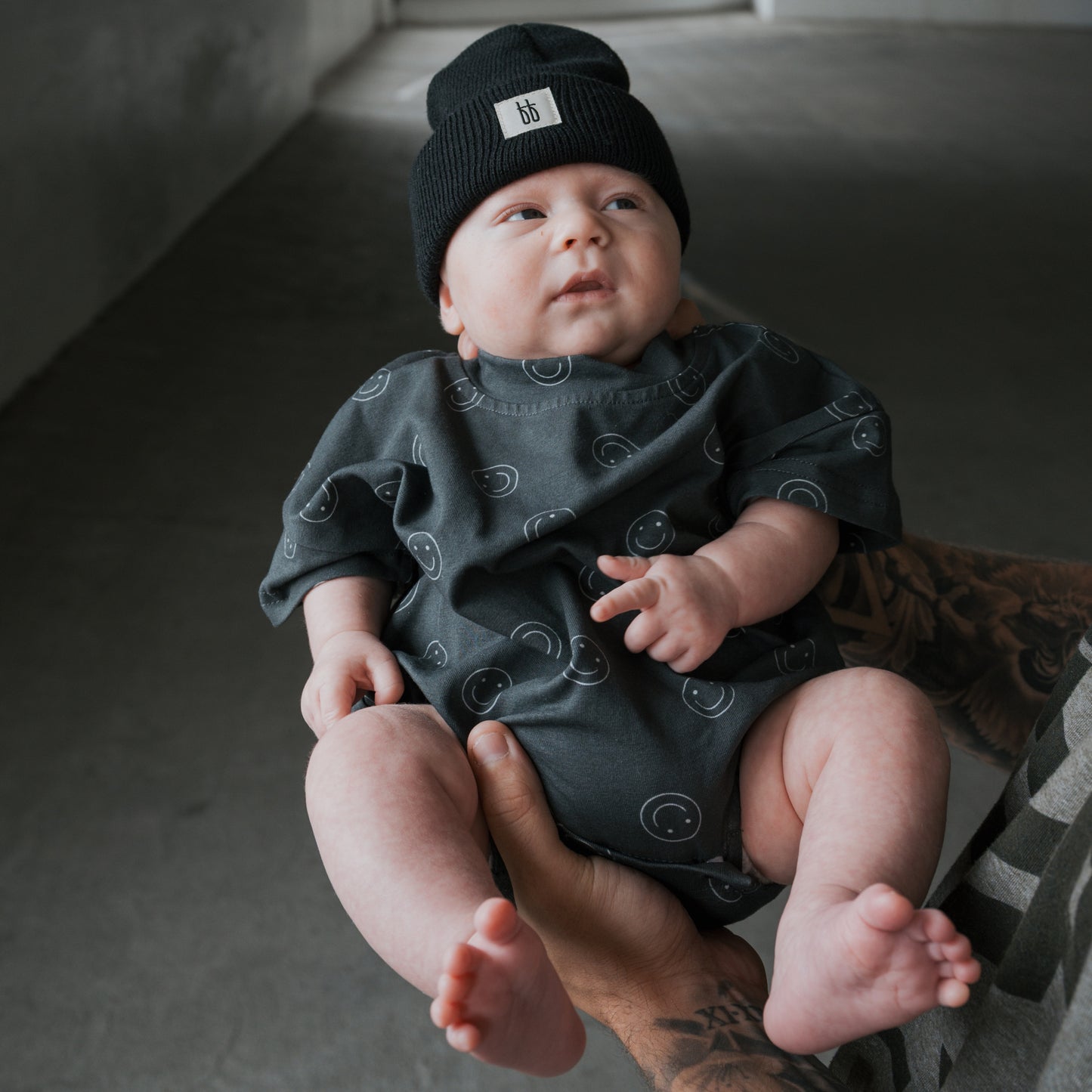 A baby wears the forever french baby Short Sleeve Romper | Smiley and a black beanie, held by a tattooed adult in a gray room.