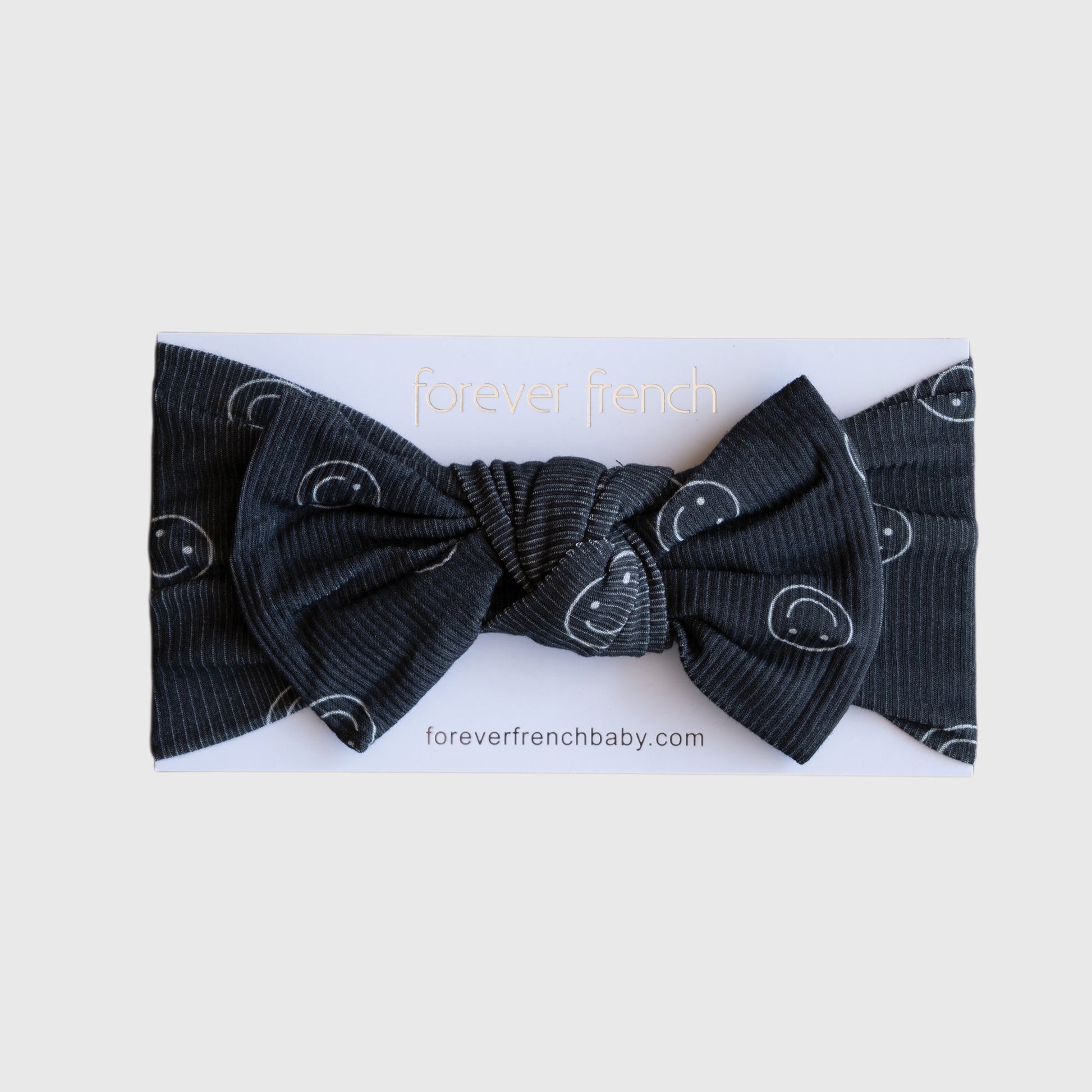 The "Bamboo Head Wrap | Smiley Ribbed" by forever french baby is a black ribbed adjustable bow headband with white smiley faces, displayed on a branded "forever french" card.