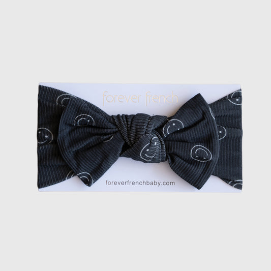The "Bamboo Head Wrap | Smiley Ribbed" by forever french baby is a black ribbed adjustable bow headband with white smiley faces, displayed on a branded "forever french" card.
