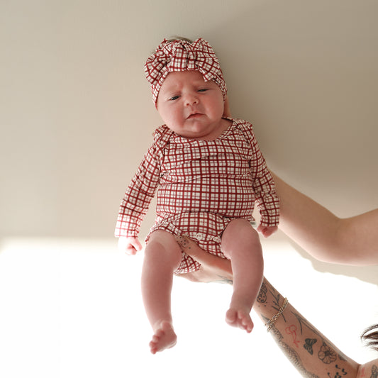 A baby wears the forever french baby Long Sleeve Bamboo Snapsuit | Window Pane with a matching headband, held by a tattooed adult against a light background.