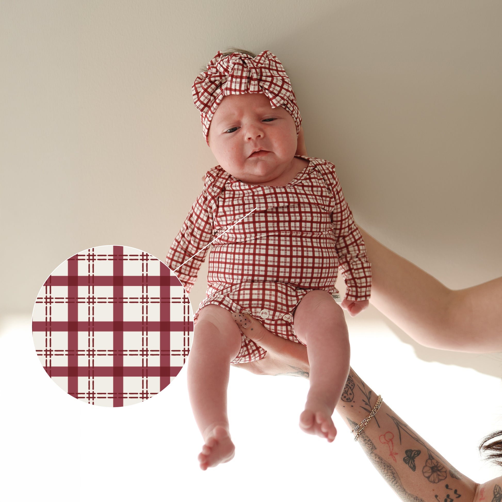 A baby in forever french baby's Long Sleeve Bamboo Snapsuit | Window Pane with a matching headband is held by tattooed arms. An inset displays a close-up of the hypoallergenic plaid pattern on this bamboo snapsuit.